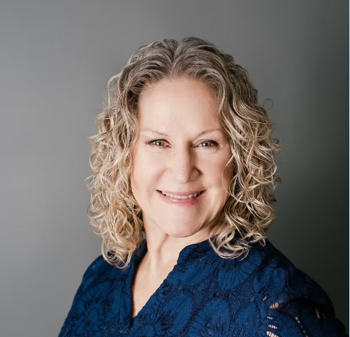 A middle-aged woman with curly blonde hair, wearing a red shirt and a silver necklace, smiling outdoors with green leafy plants and a brick wall in the background.
