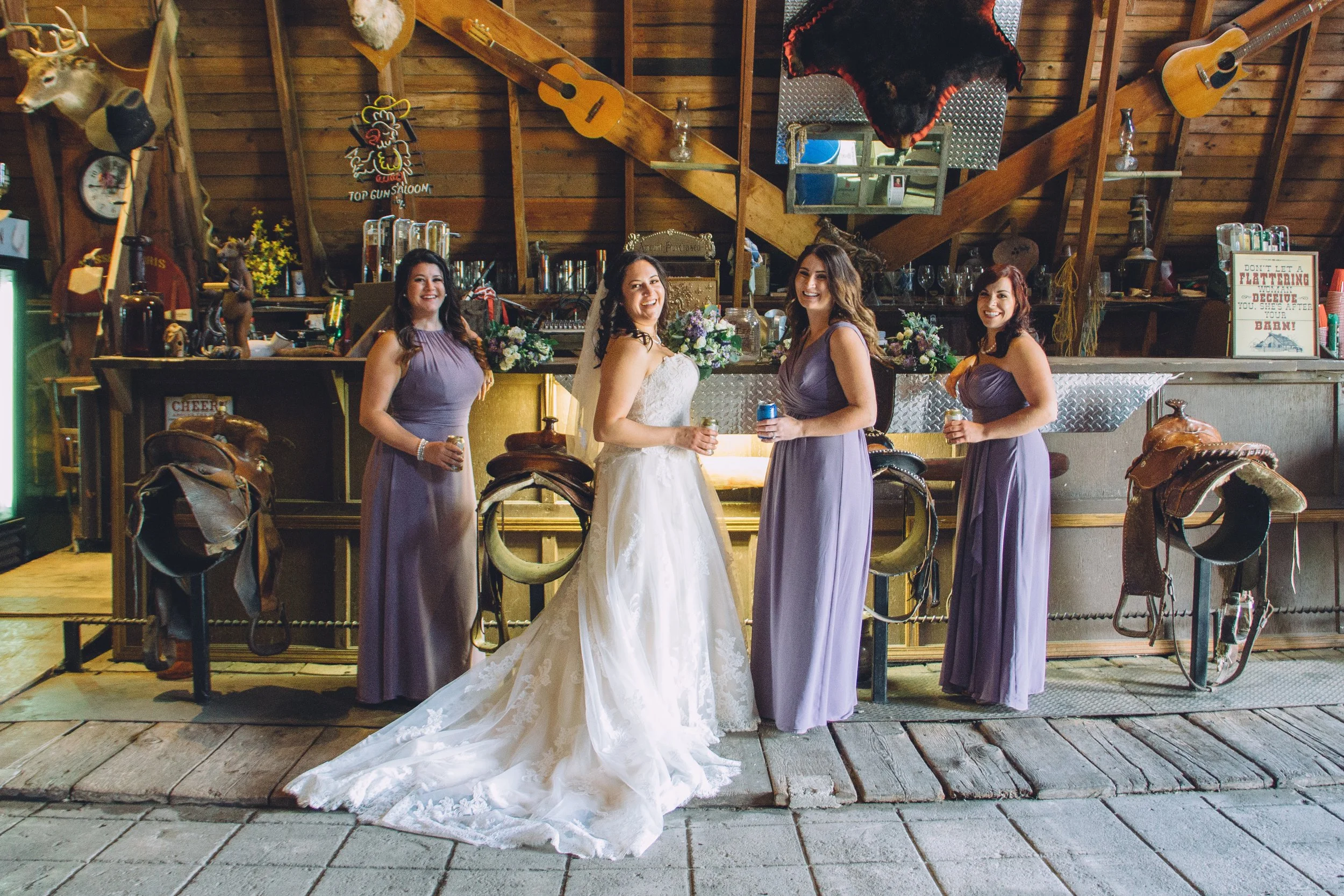 Bride with bridesmaids in lavender dresses standing in front of the rustic bar and saddle barstools.