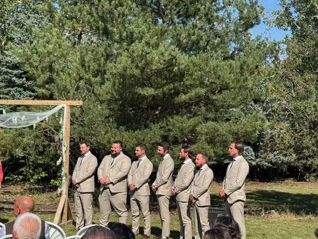 Groomsmen standing beside the wooden ceremony arch during the wedding ceremony.
