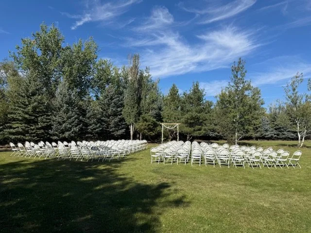 Outdoor ceremony setup with wooden archway, white chairs and blue prairie skies.