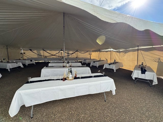 Empty event tent with long tables set with white tablecloths, bottles, and decorations, awaiting guests.