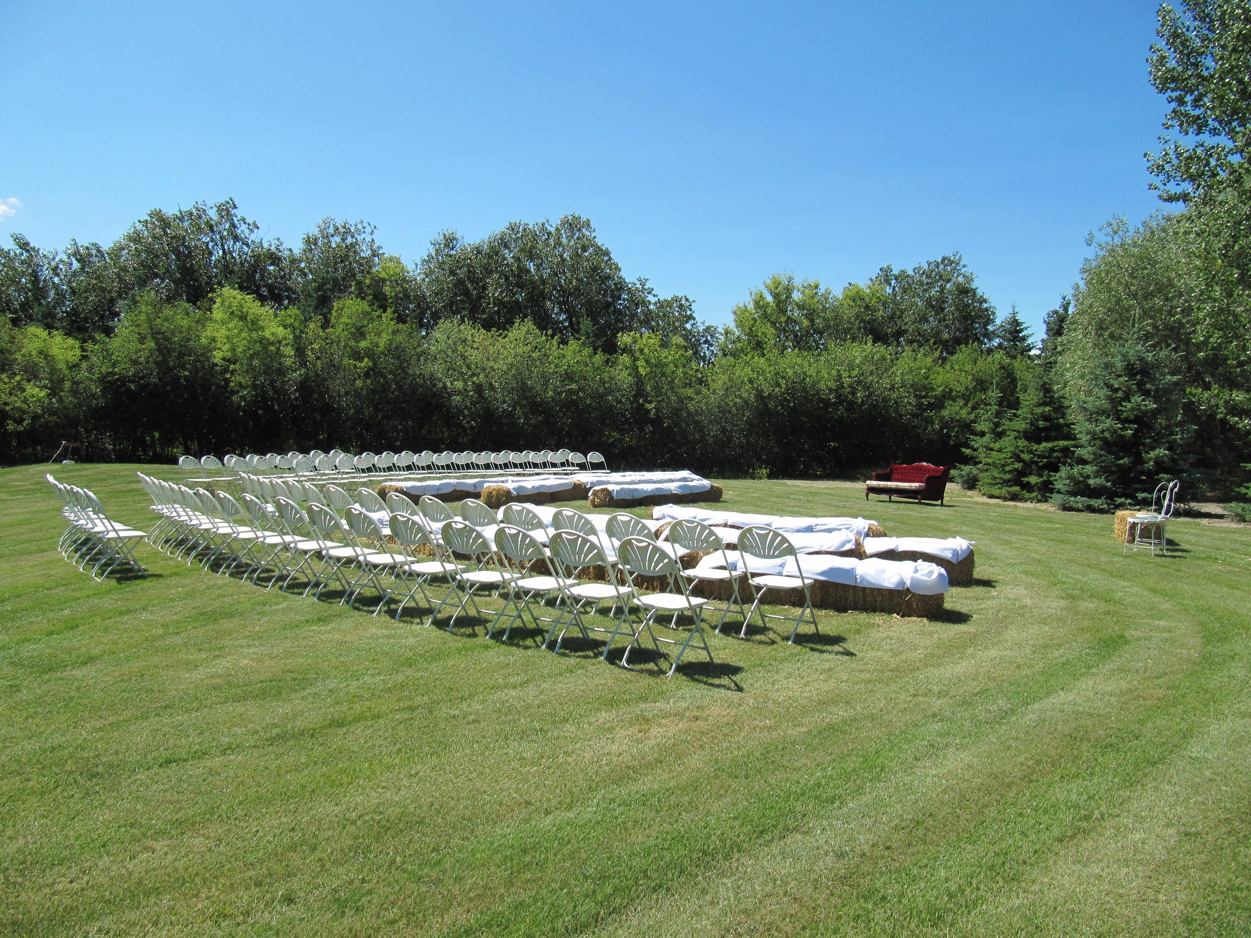 Outdoor event space with white seating surrounded by mature trees and green lawn.