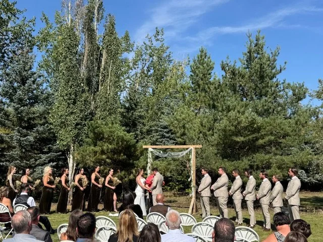 Bride and groom with their wedding party standing beneath a wooden arch while guests watch.