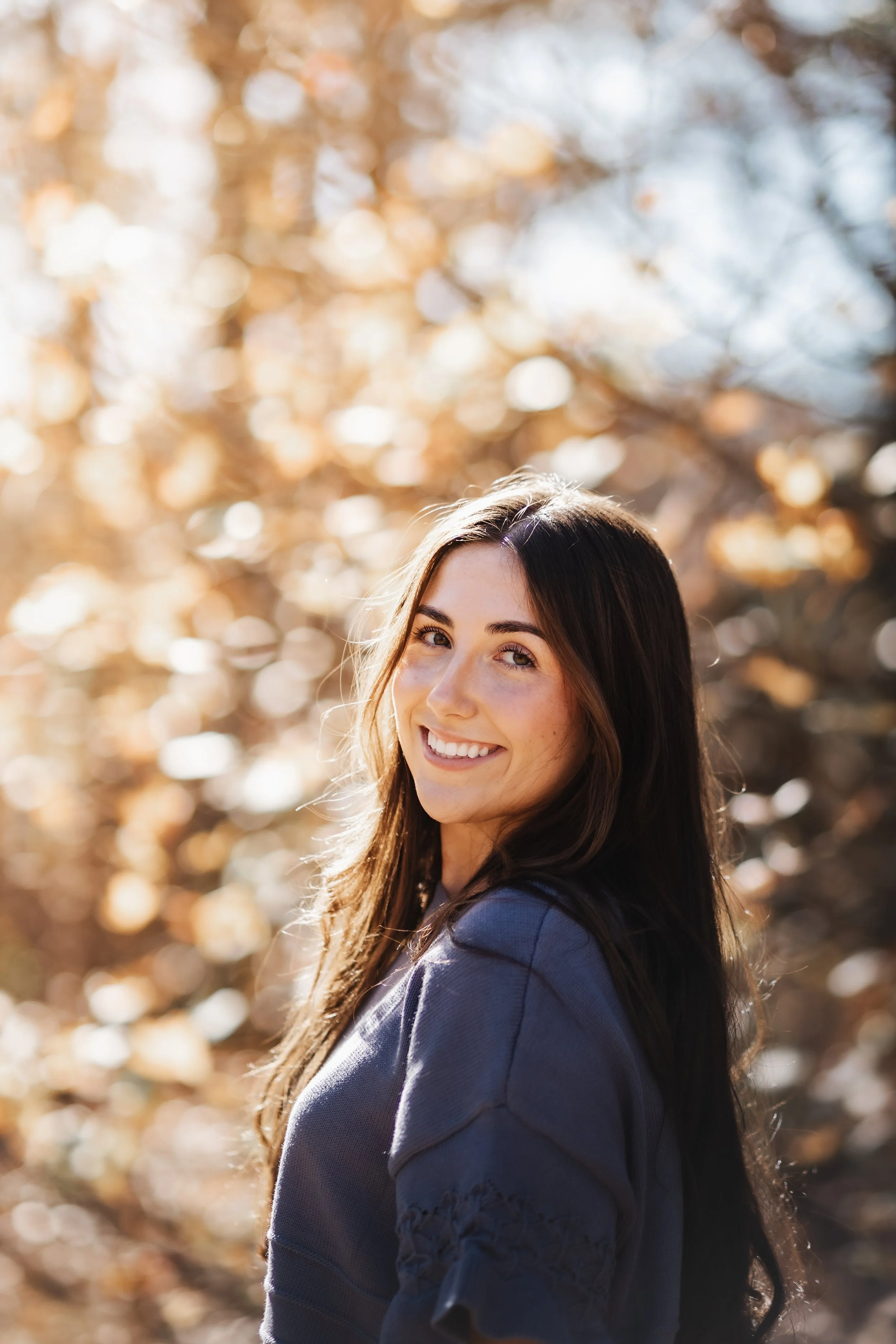 A young woman with long dark hair smiling outdoors with autumn foliage in the background.
