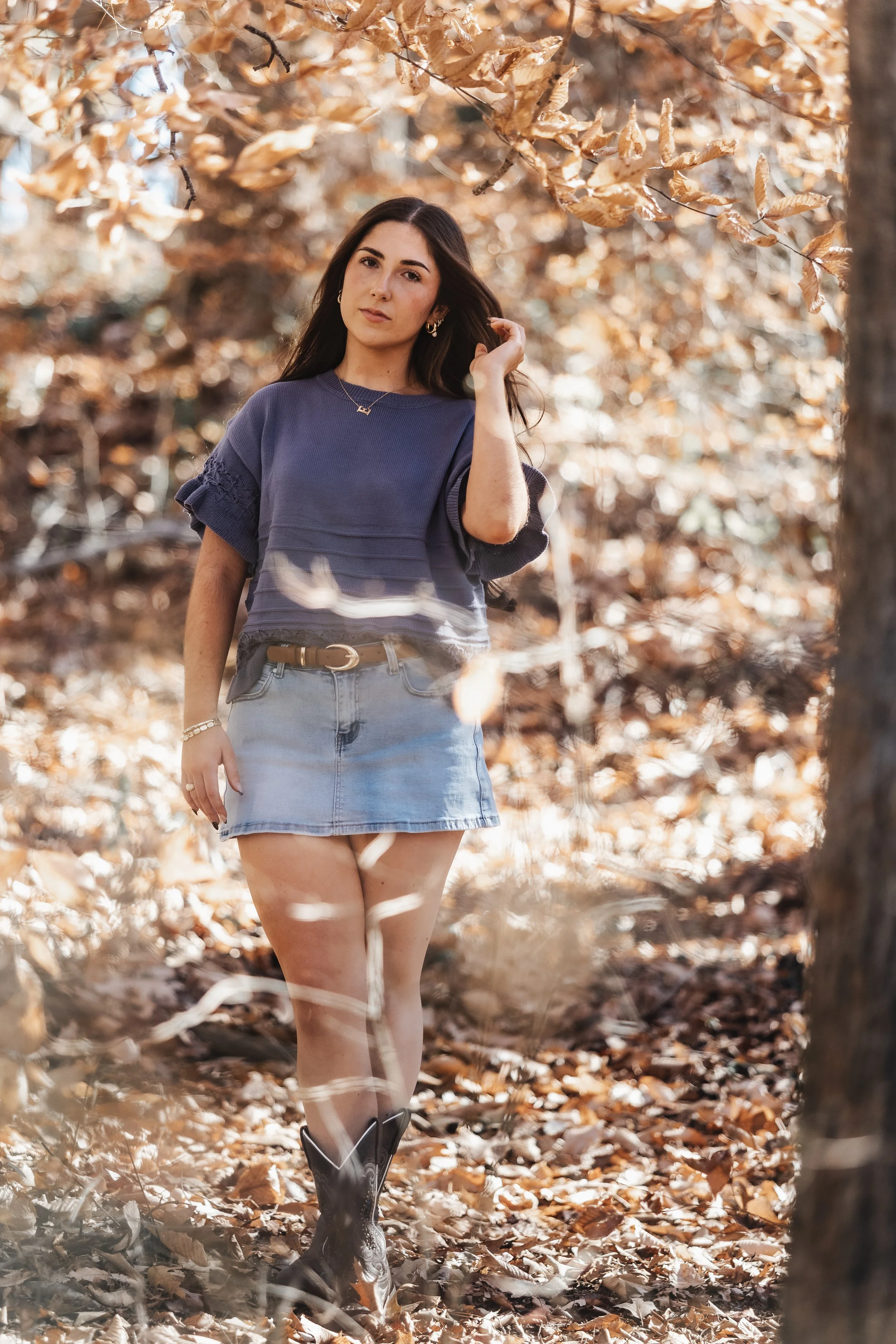 A young woman standing outdoors in a forest during autumn, wearing a navy blue top, a denim mini skirt, and cowboy boots, with fallen leaves on the ground and trees with brown leaves around her.