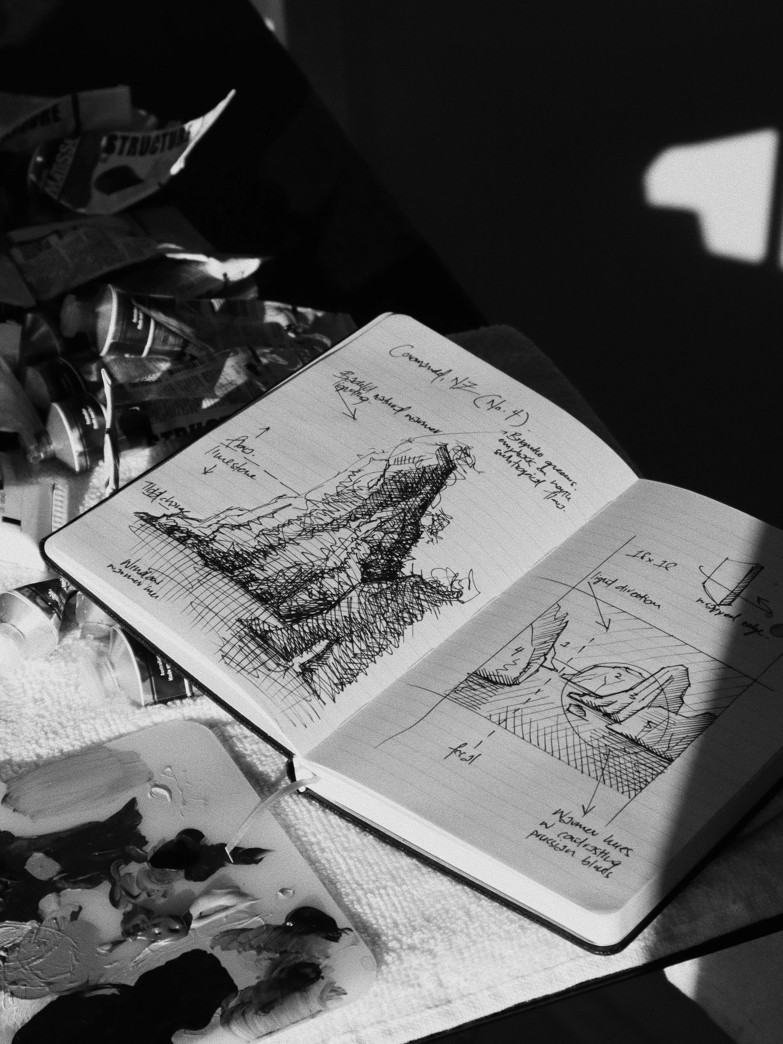 A sketchbook with two page drawings of towers or buildings, with annotations and sketches, surrounded by paint tubes and a painter's palette, on a table with scattered art supplies.