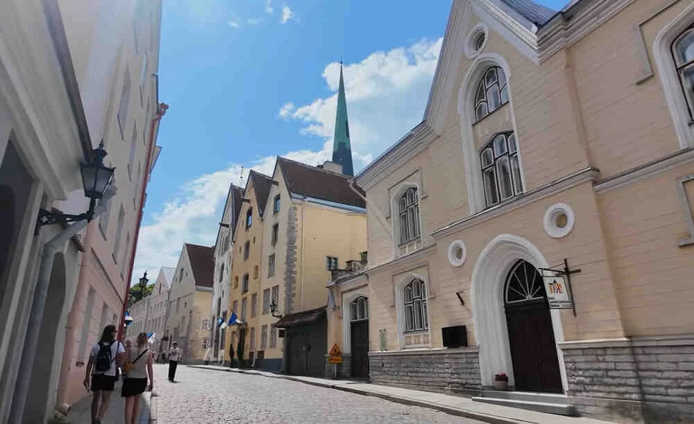 Pedestrians walking along a narrow cobblestone street in Tallinn Old Town, Estonia, lined with historic pastel-colored buildings and a church spire rising in the background under a bright uncovered clear blue sky