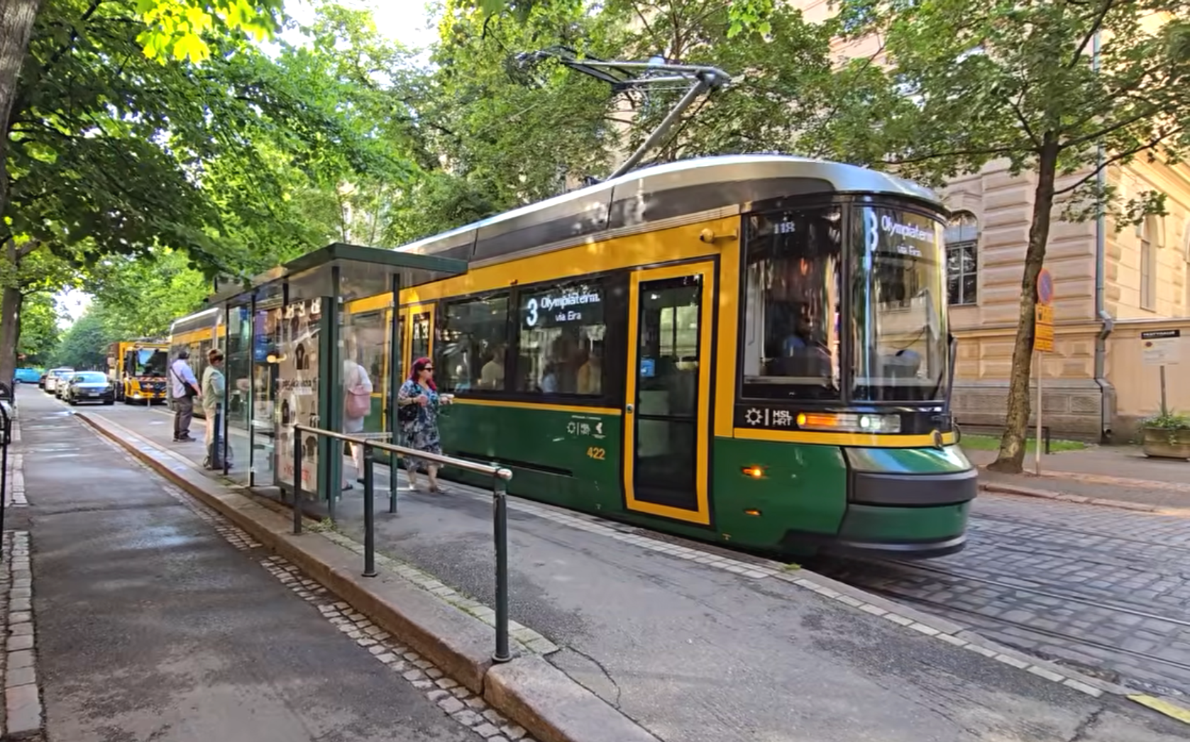 Green and yellow Helsinki tram stopped at a tree-lined street station with passengers waiting nearby.