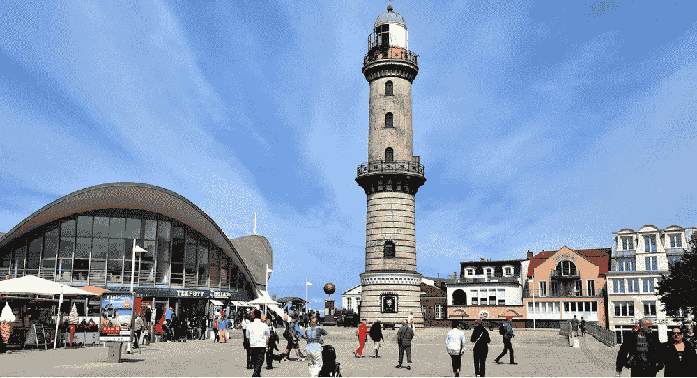 Warnemünde Lighthouse beside the Teepott building with visitors walking through the seaside promenade in Warnemünde, Germany.