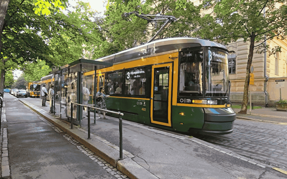 Green and yellow Helsinki tram stopped at a tree-lined street station with passengers waiting nearby.