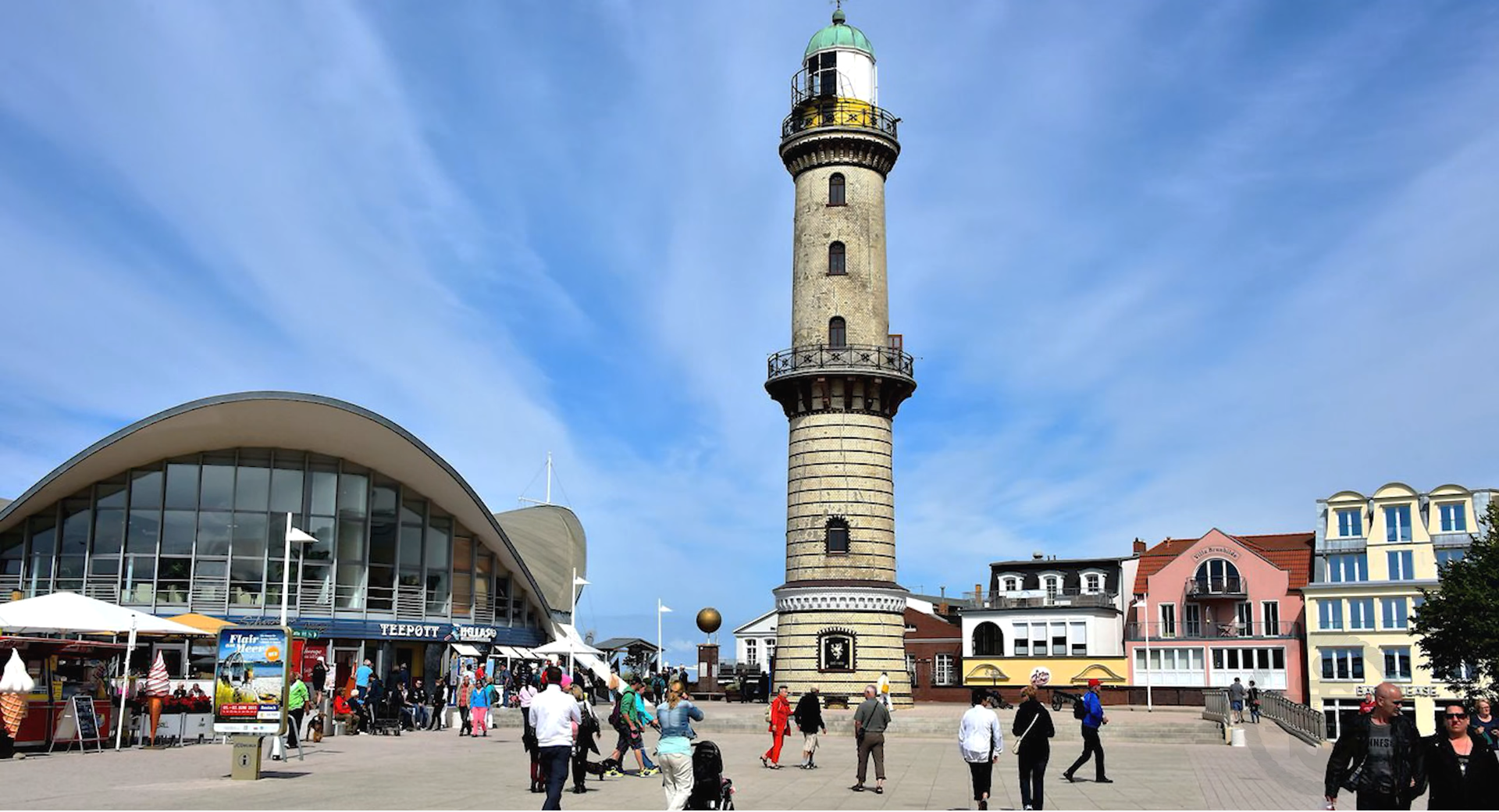 Warnemünde Lighthouse beside the Teepott building with visitors walking through the seaside promenade in Warnemünde, Germany.