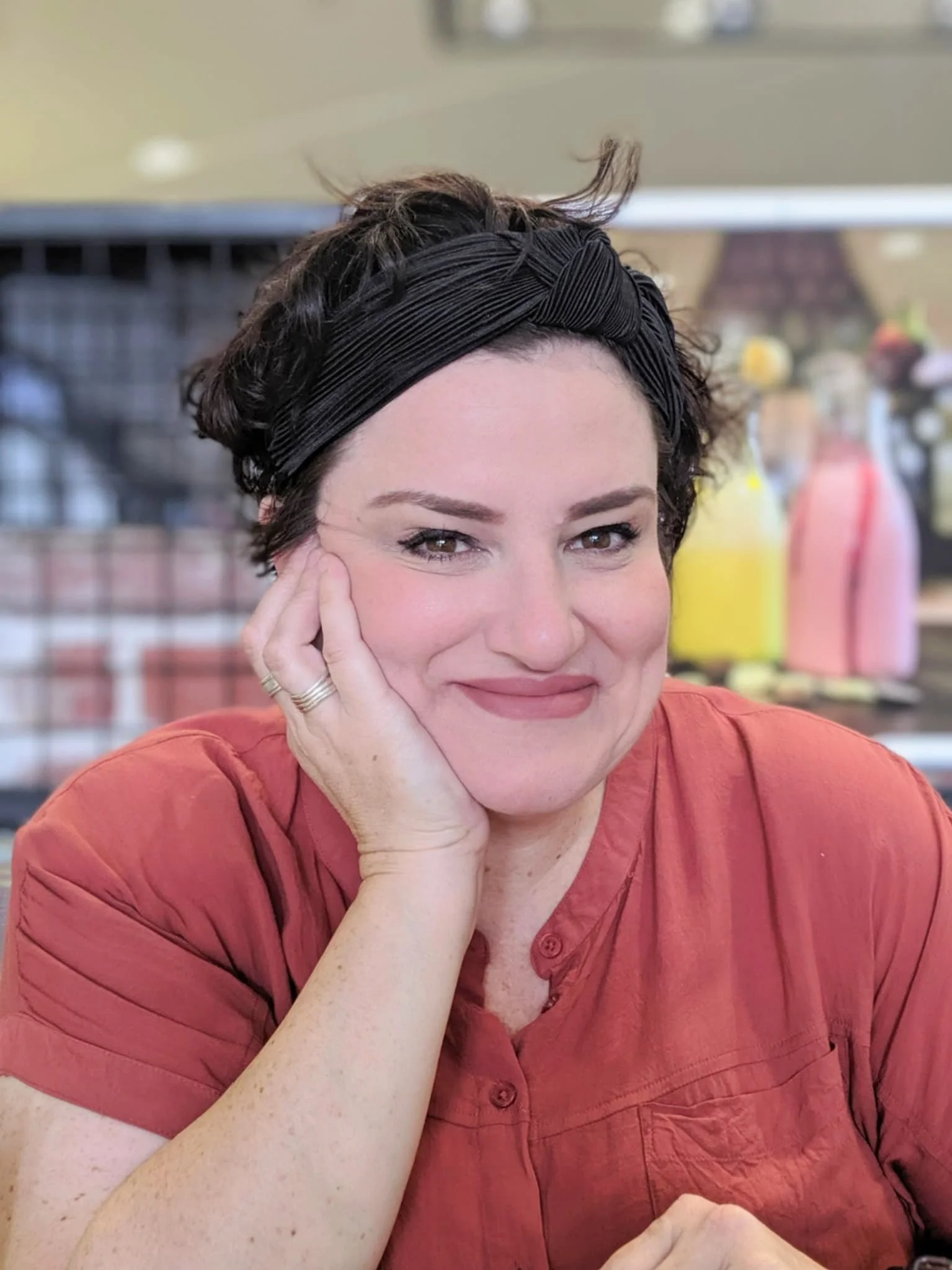 A woman with short dark hair, wearing a black headband, resting her face on her hand, and smiling softly. She is wearing a reddish-orange shirt and is in a store or cafe with colorful bottles and blurred background.