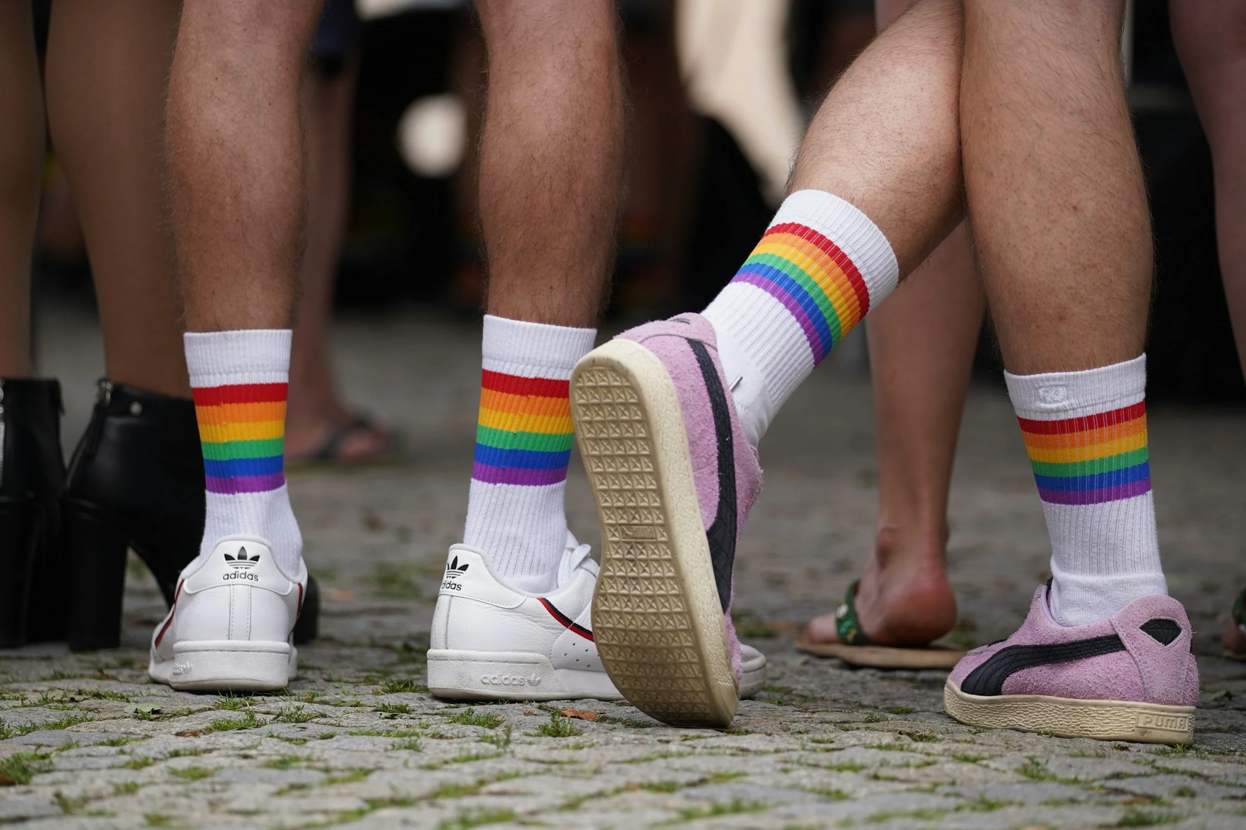 Close-up of people wearing rainbow-striped socks and sneakers, kneeling and standing on cobblestone pavement.