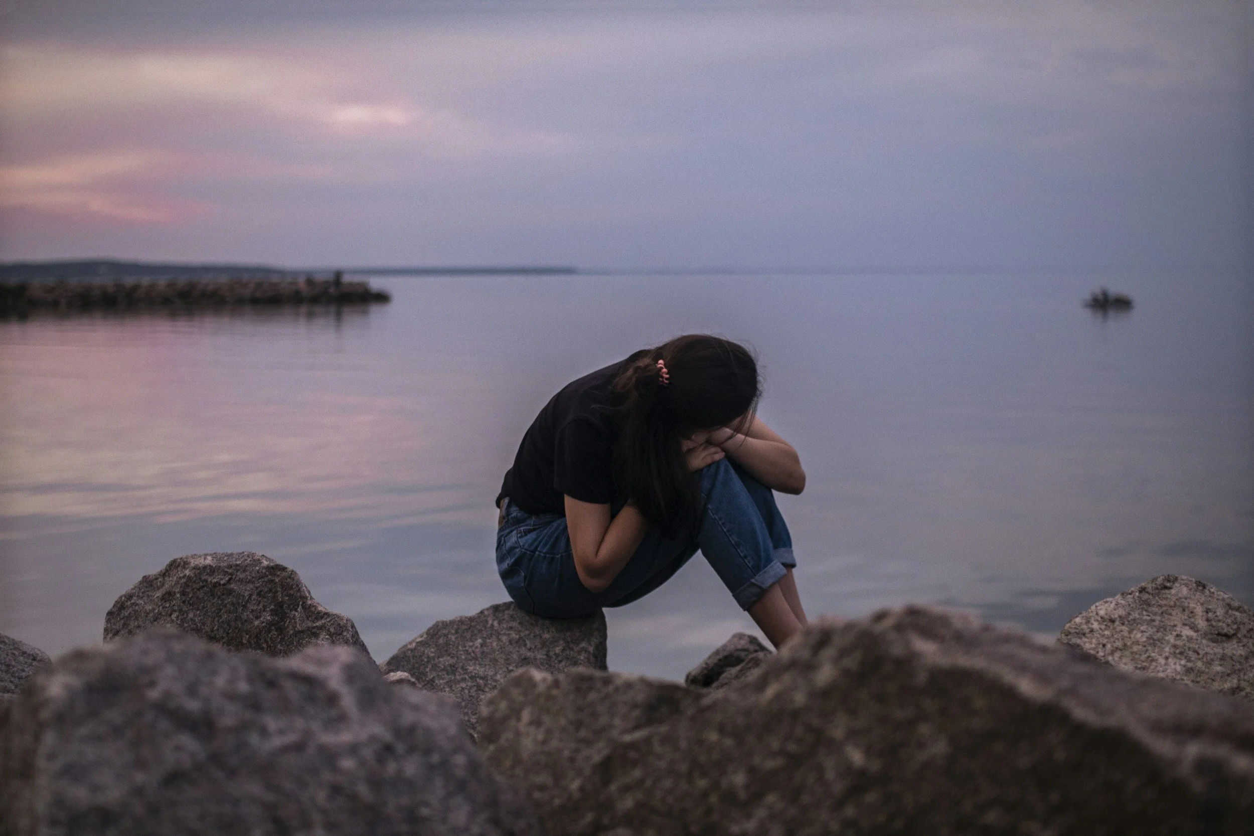 A person sitting on rocks by calm water at sunset, with one arm resting on their knee and head bowed.