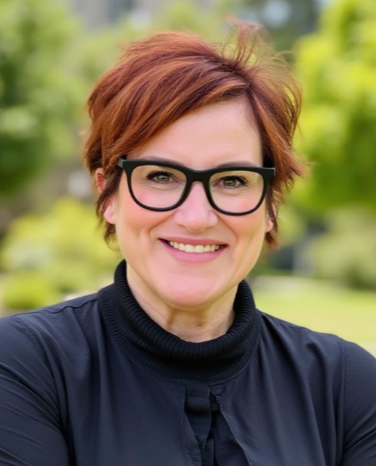 A woman with short red hair, black glasses, and a black turtleneck smiling outdoors with blurred green trees in the background.