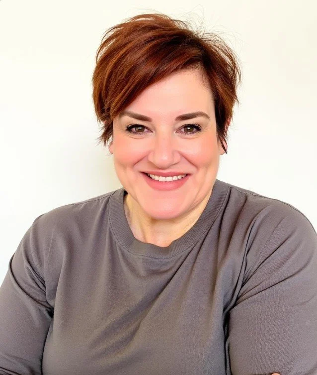 A woman with short, reddish-brown hair smiling at the camera, wearing a gray long-sleeve shirt, against a plain light background.