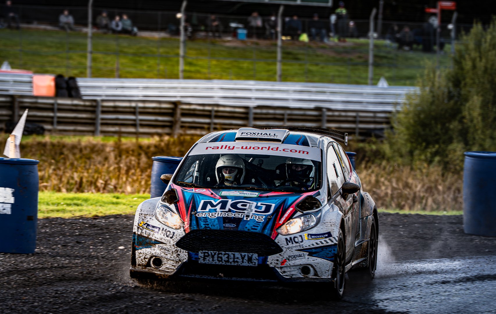 Rally car racing on a muddy track with spectators watching from the background.