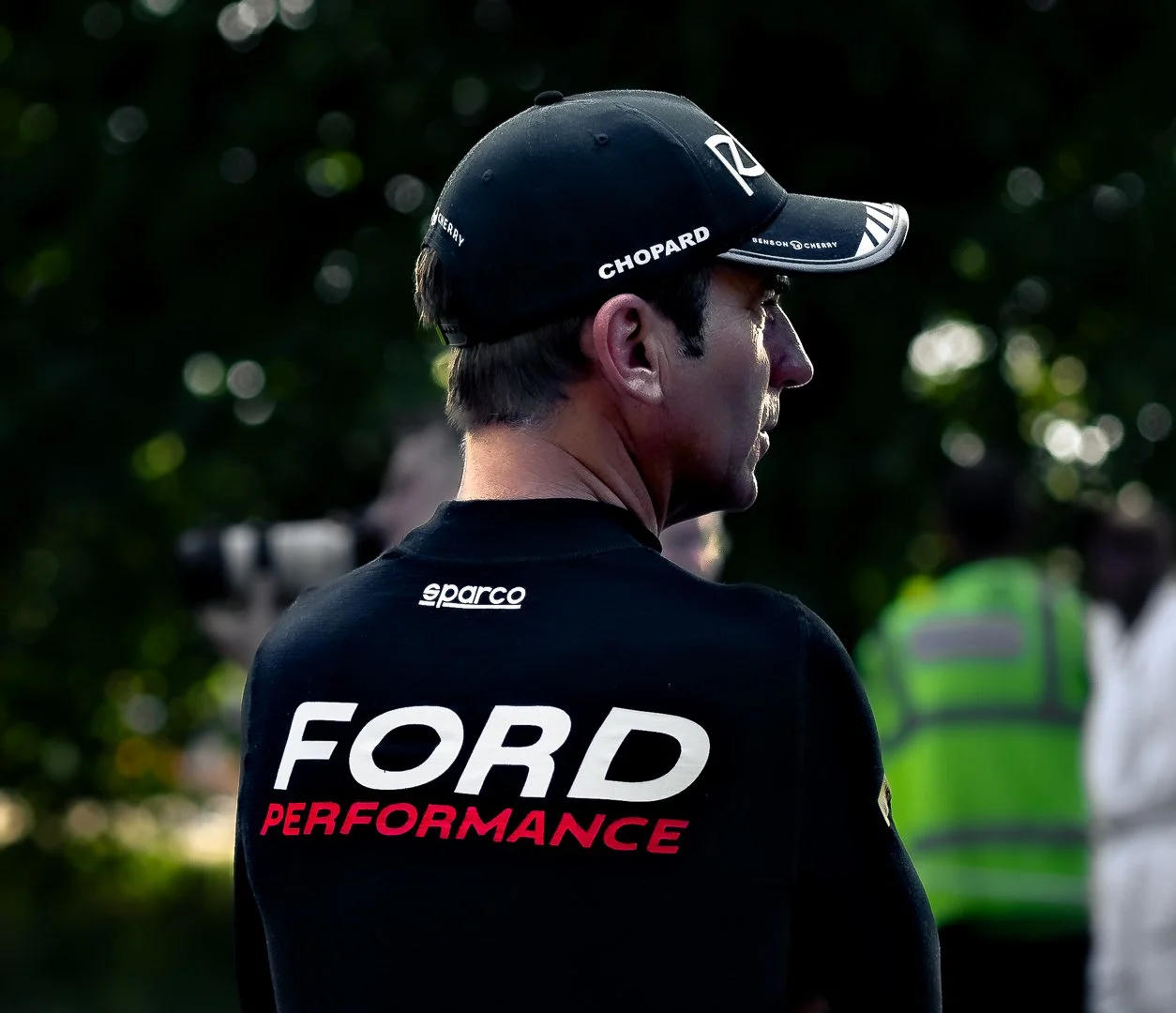 A man wearing a black cap and a black shirt with 'FORD PERFORMANCE' written on the back, standing outdoors with a blurry green background.