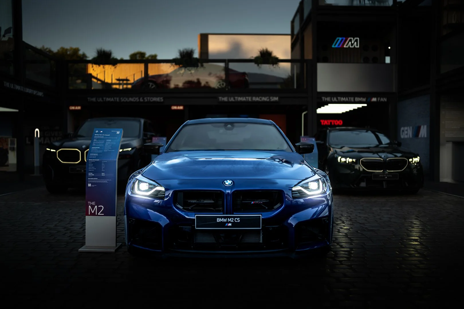 A blue BMW M2 CS sports car on display at an auto show, with other BMW models in the background.