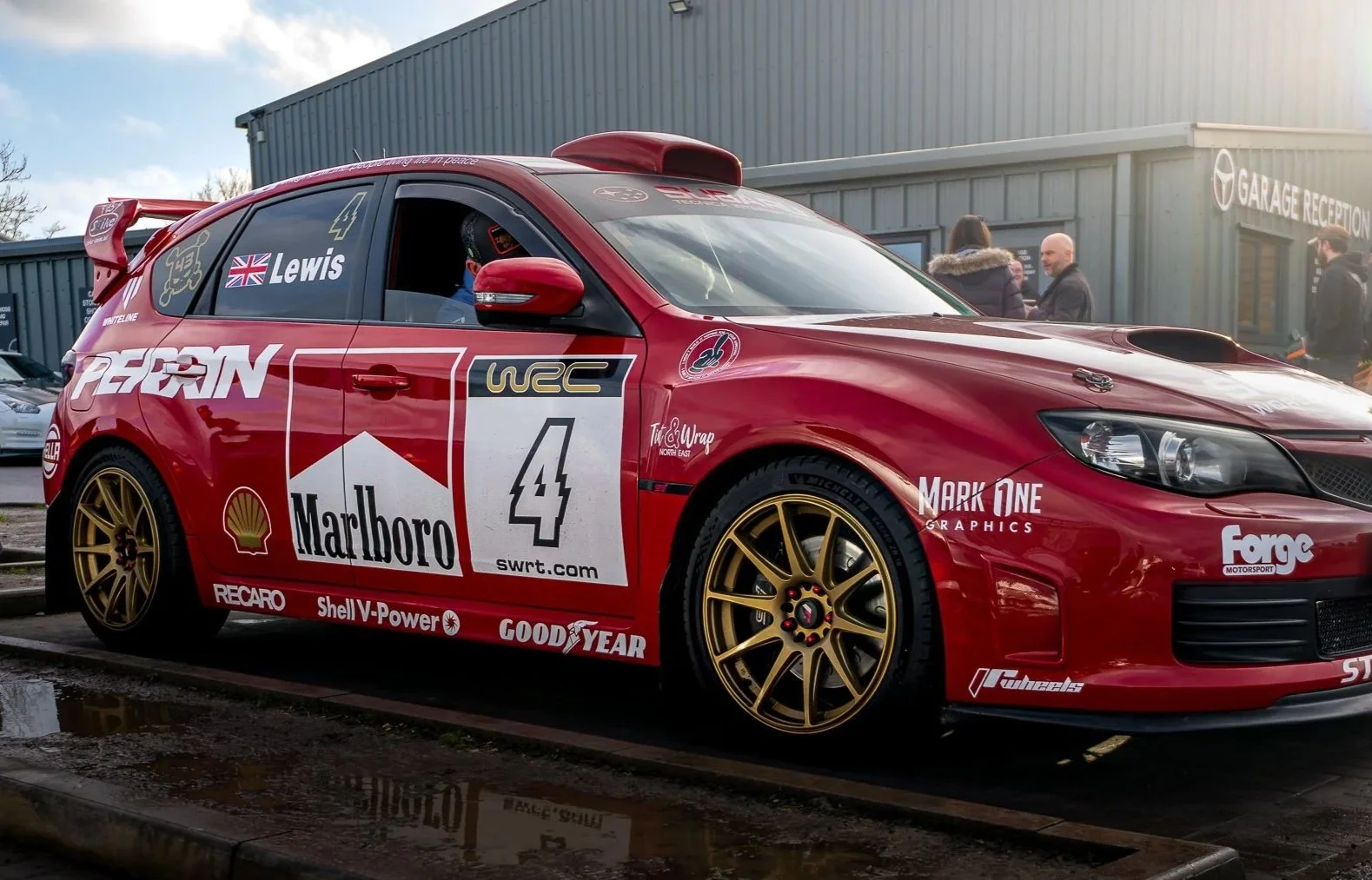Red rally car with gold wheels parked at a garage, with several people in the background, and various sponsor logos including Marlboro, Shell, Goodyear, and PERGIN.