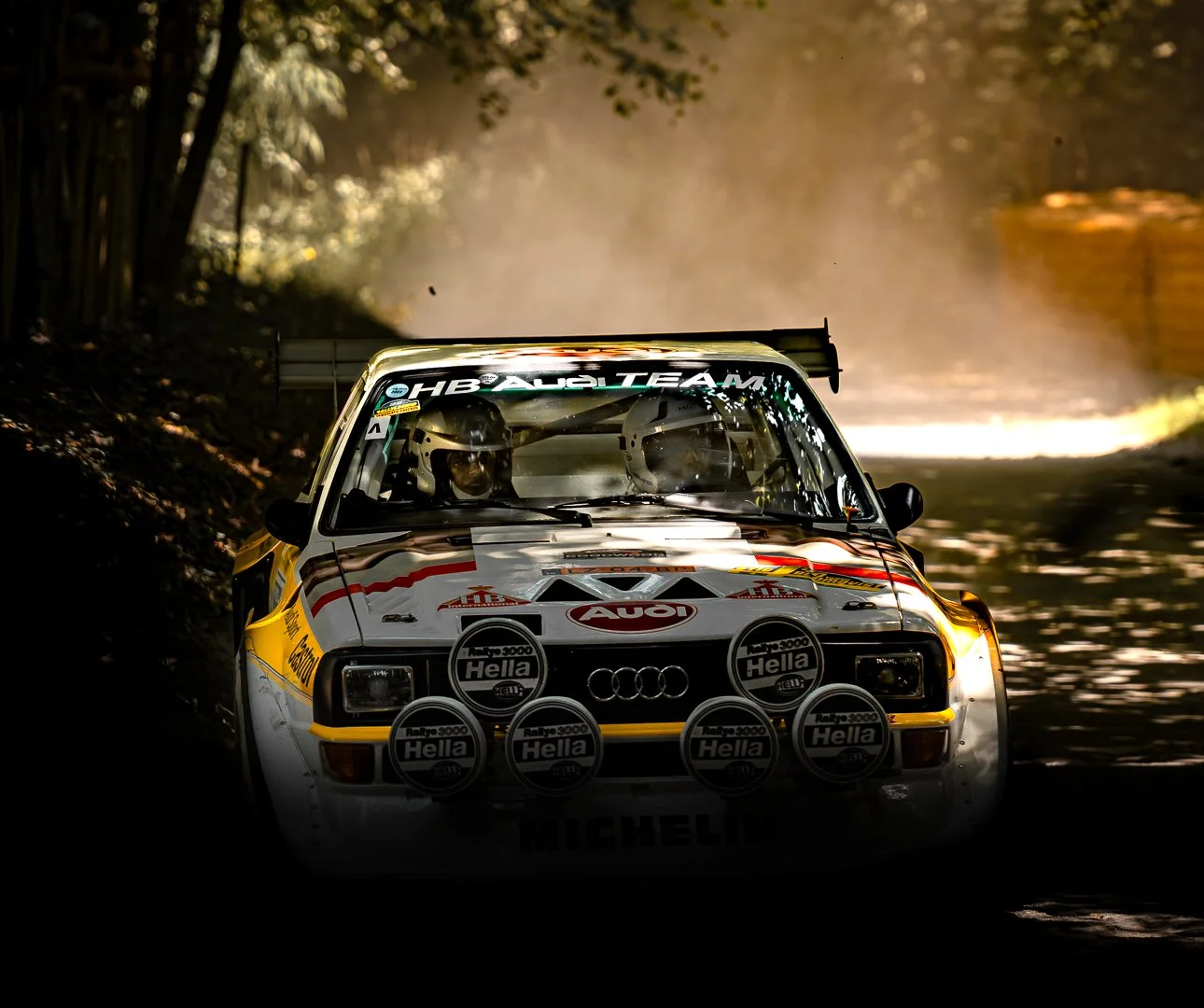 A vintage rally car with prominent headlights and racing decals, driving on a sunlit forest road, dust and trees in the background.
