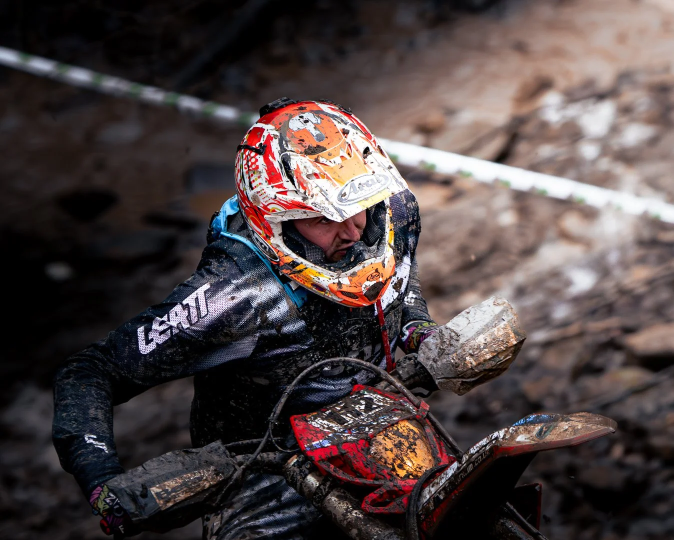 Person wearing a colorful motocross helmet and gear, riding a dirt bike through muddy terrain.