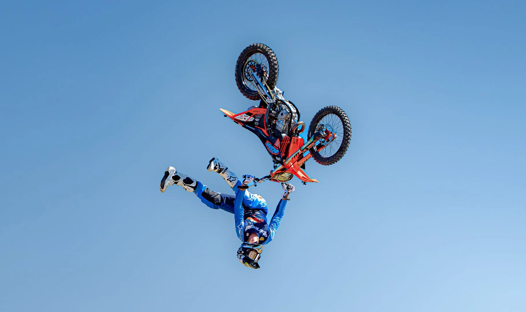 A motocross rider flips upside down in mid-air with a red and black dirt bike against a clear blue sky.