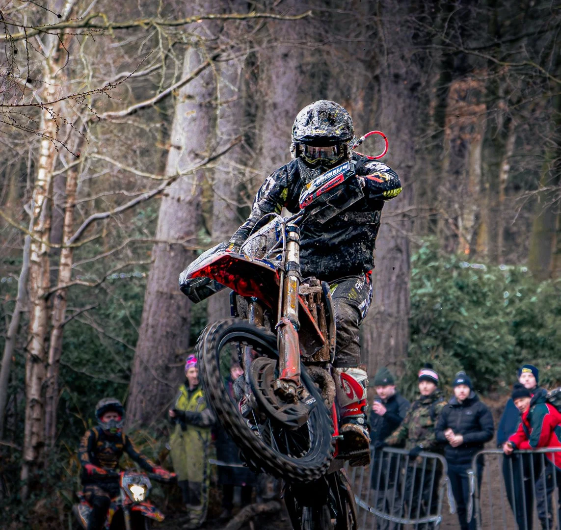A dirt bike rider in mid-air jump during a forest race, with spectators watching behind safety barriers.
