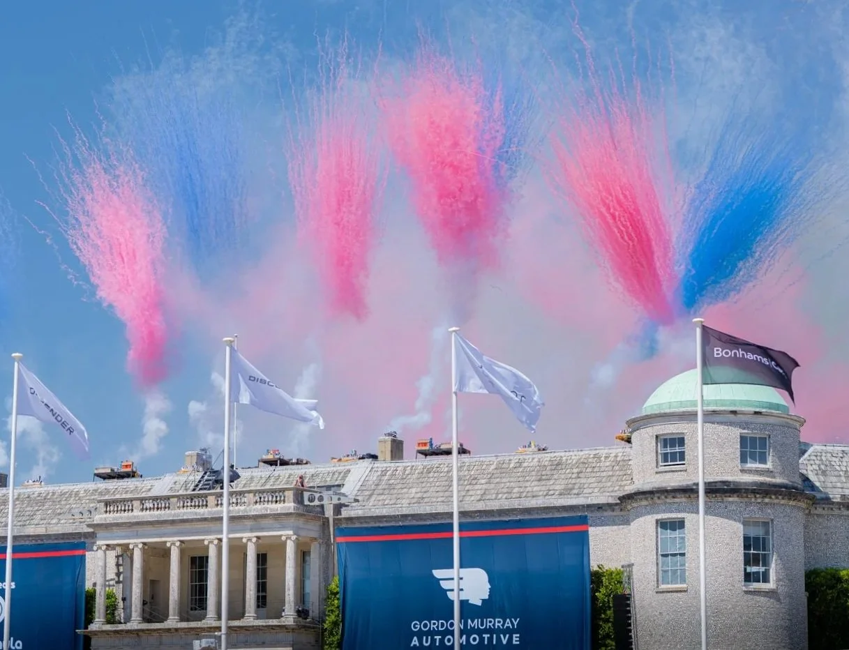 Colorful pink and blue smoke bursts in the sky above a building with flags and banners displaying the Gordon Murray Automotive logo and text, with a clear blue sky background.