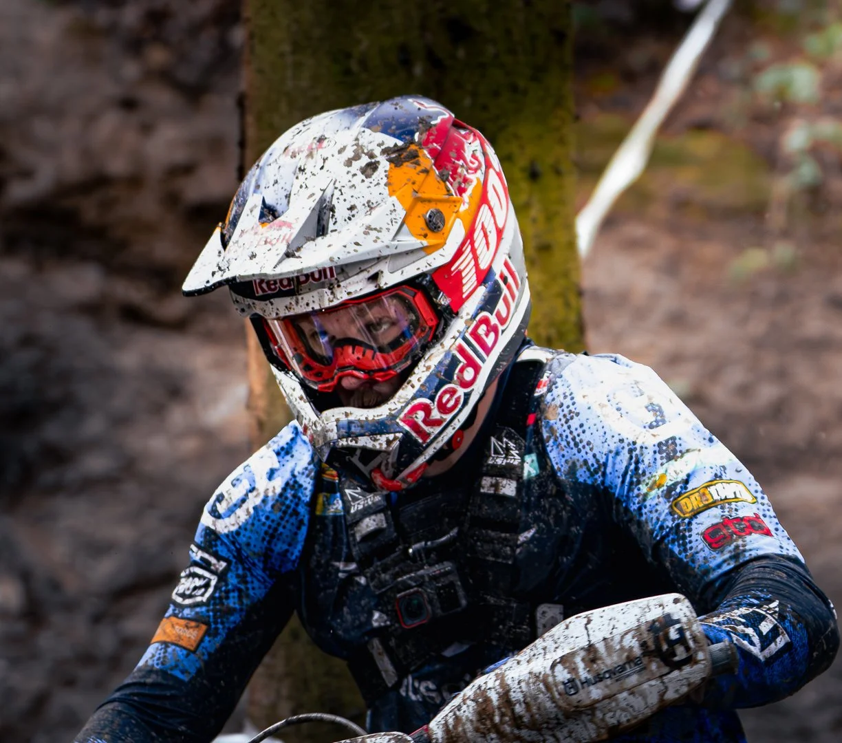 An off-road motorcycle rider in a muddy helmet and gear riding through a forest trail.