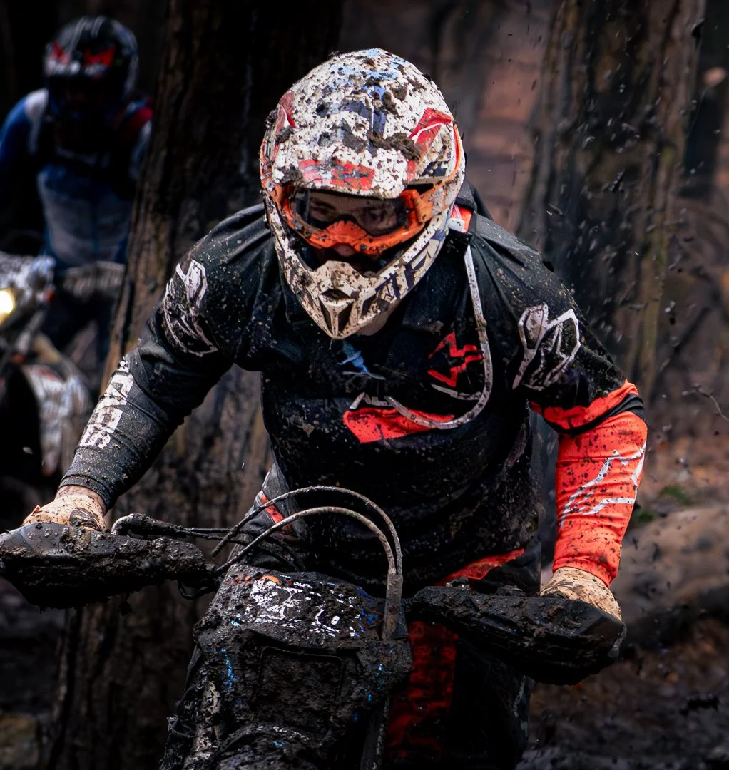 Motocross rider in black and red gear riding through muddy terrain, covered in mud and dirt, with a forest background.
