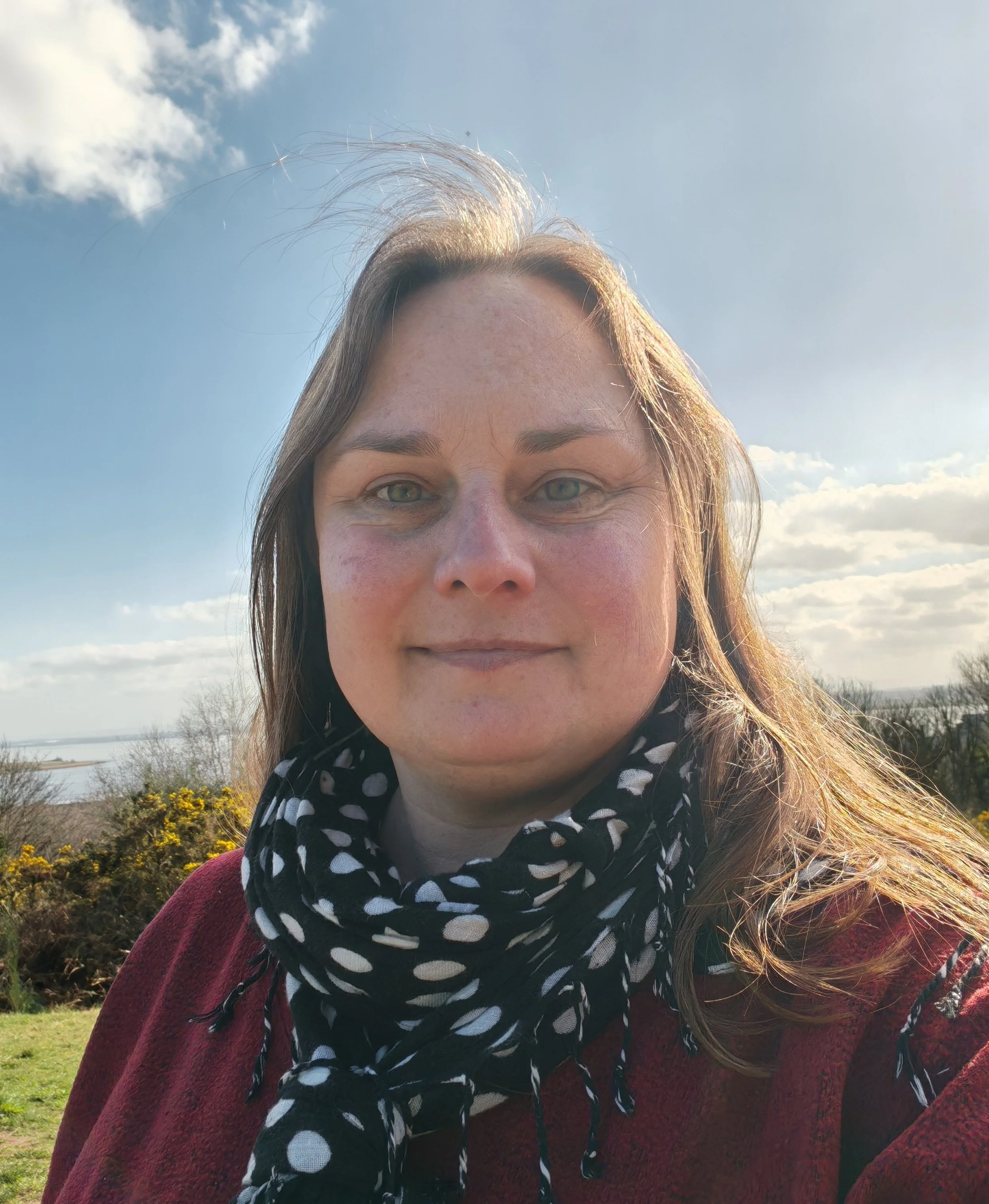 Selfie of Janine Keeble in a black and white spotty scarf and red jumper against a blue sky and heathland.