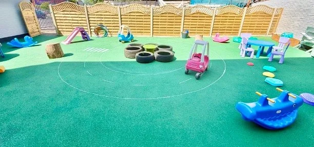 Children's playground with colorful toys including a pink ride-on car, blue rocking horse, tire obstacle, and various small chairs and slides on green turf, enclosed by wooden fencing.