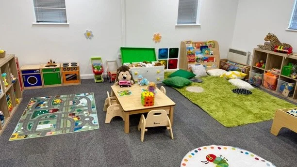 A colorful children's playroom with toys, plush animals, a green rug, a small table with chairs, and educational and play supplies along the walls.