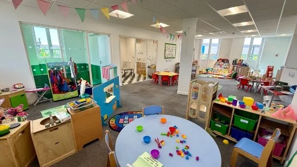 Bright and colorful indoor preschool or daycare classroom with play areas, tables with toys, and educational materials.