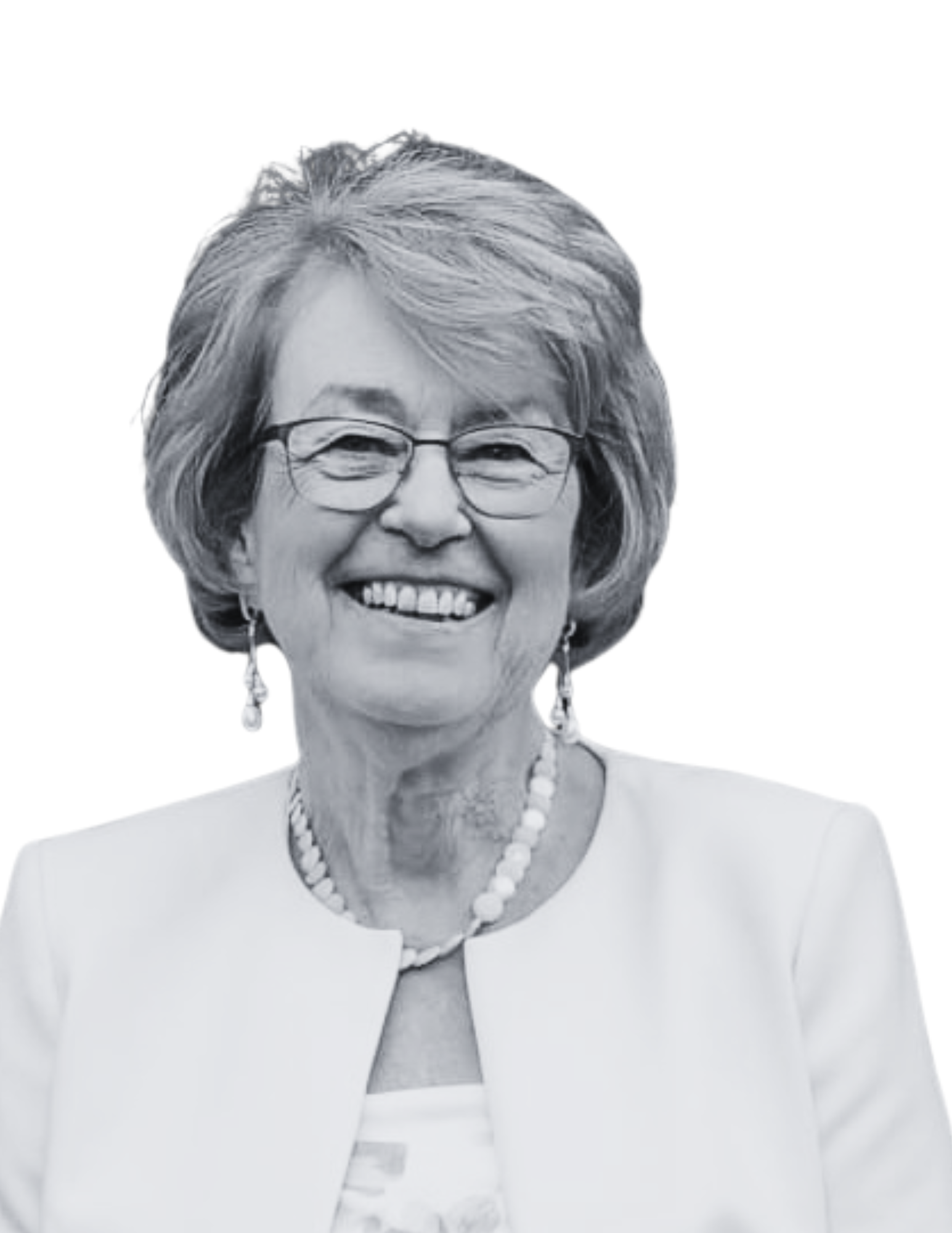 A smiling elderly woman with glasses, earrings, and a necklace, wearing a light-coloured top and jacket.