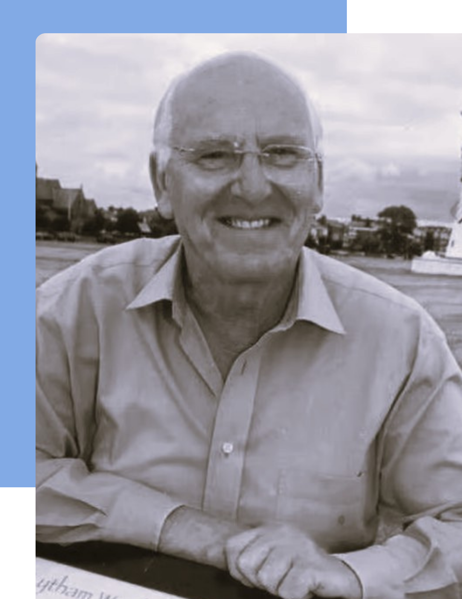 Black and white head and shoulders images of a smiling older white man with a bald head and glasses. He is wearing a open collar long sleeve shirt. Lytham Green is in the background just visible.