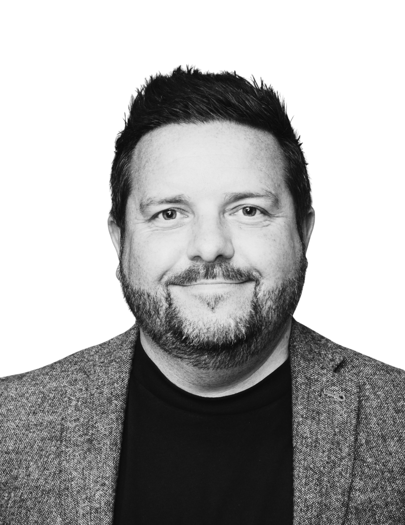 Black and white portrait of a man with short dark hair, light skin, and a beard, smiling, wearing a blazer over a dark shirt.