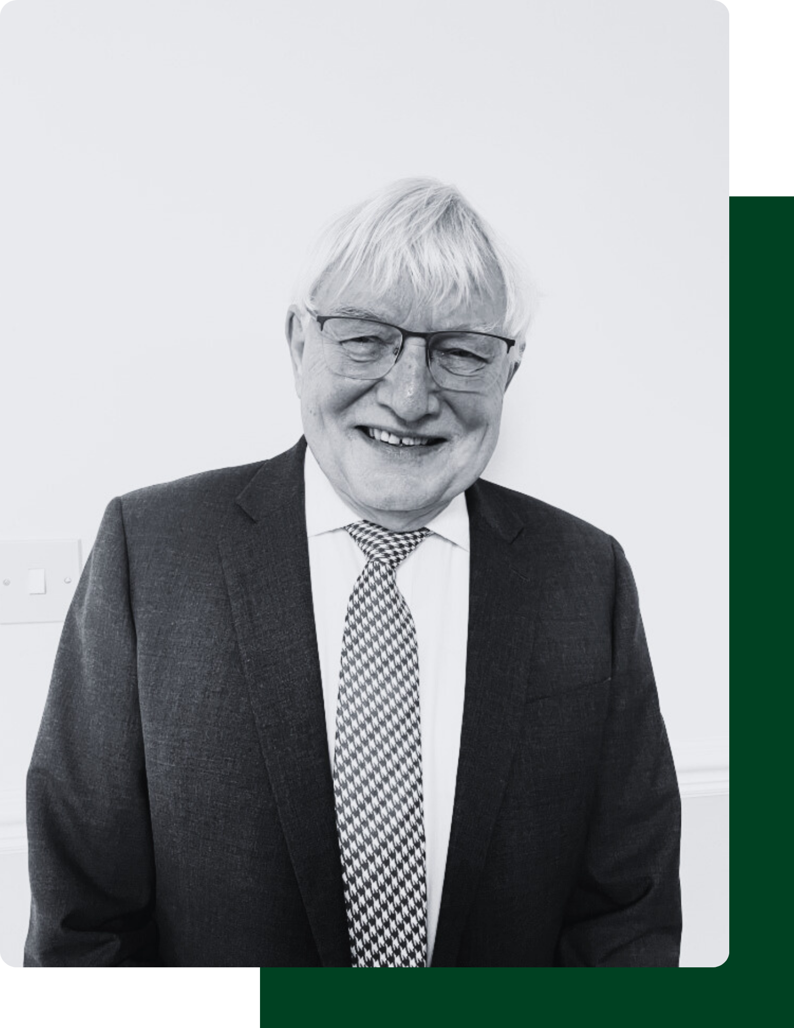 Black and white image of a head and shoulders shot of a smiling older white man with white hair and glasses. He is wearing a dark suit white shirt and check pattern tie and he is standing against a plain pale background.