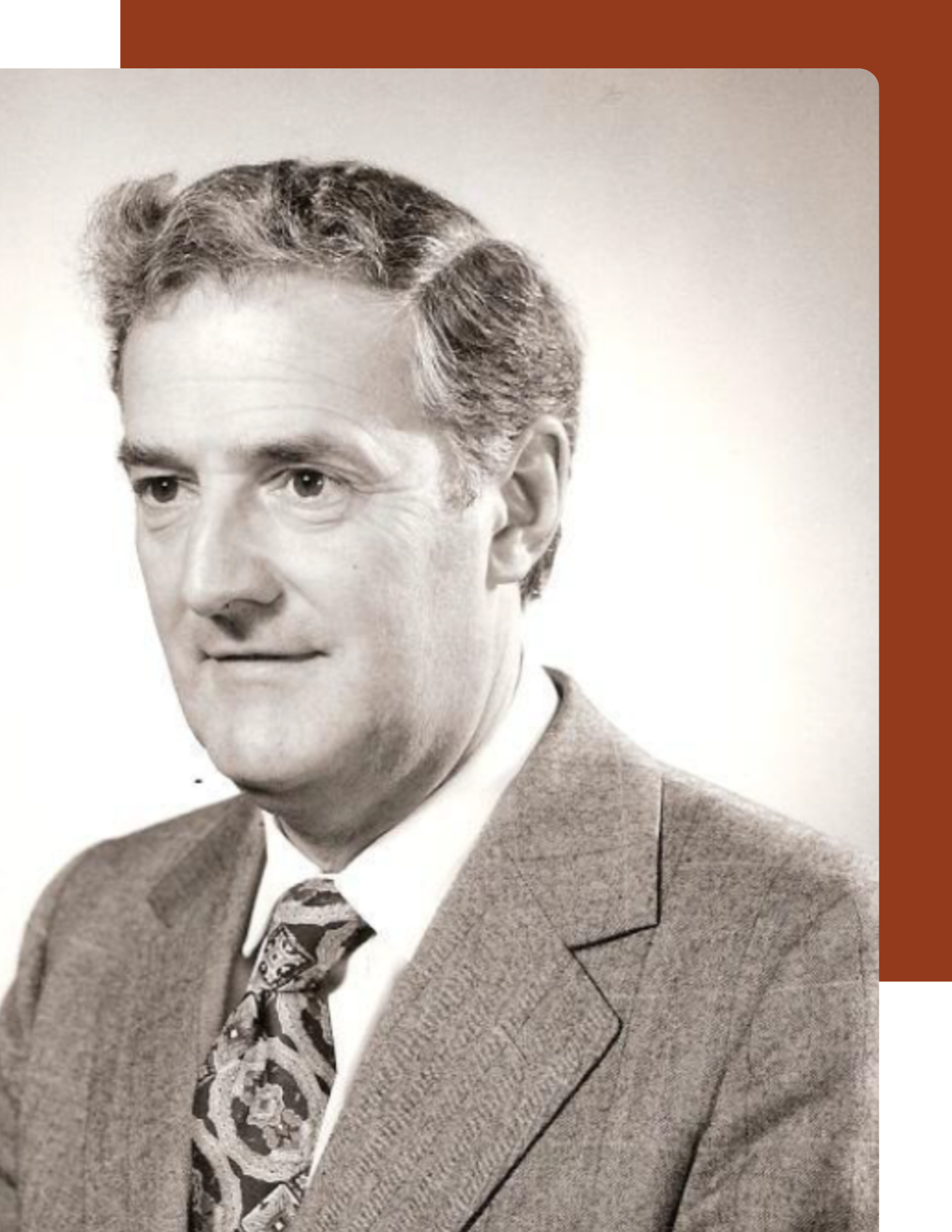 Black and white head and shoulders profile image of an older white man with brown hair. He is wearing a grey suit, white shirt and patterned tie. He is set against a pale vingetted background.