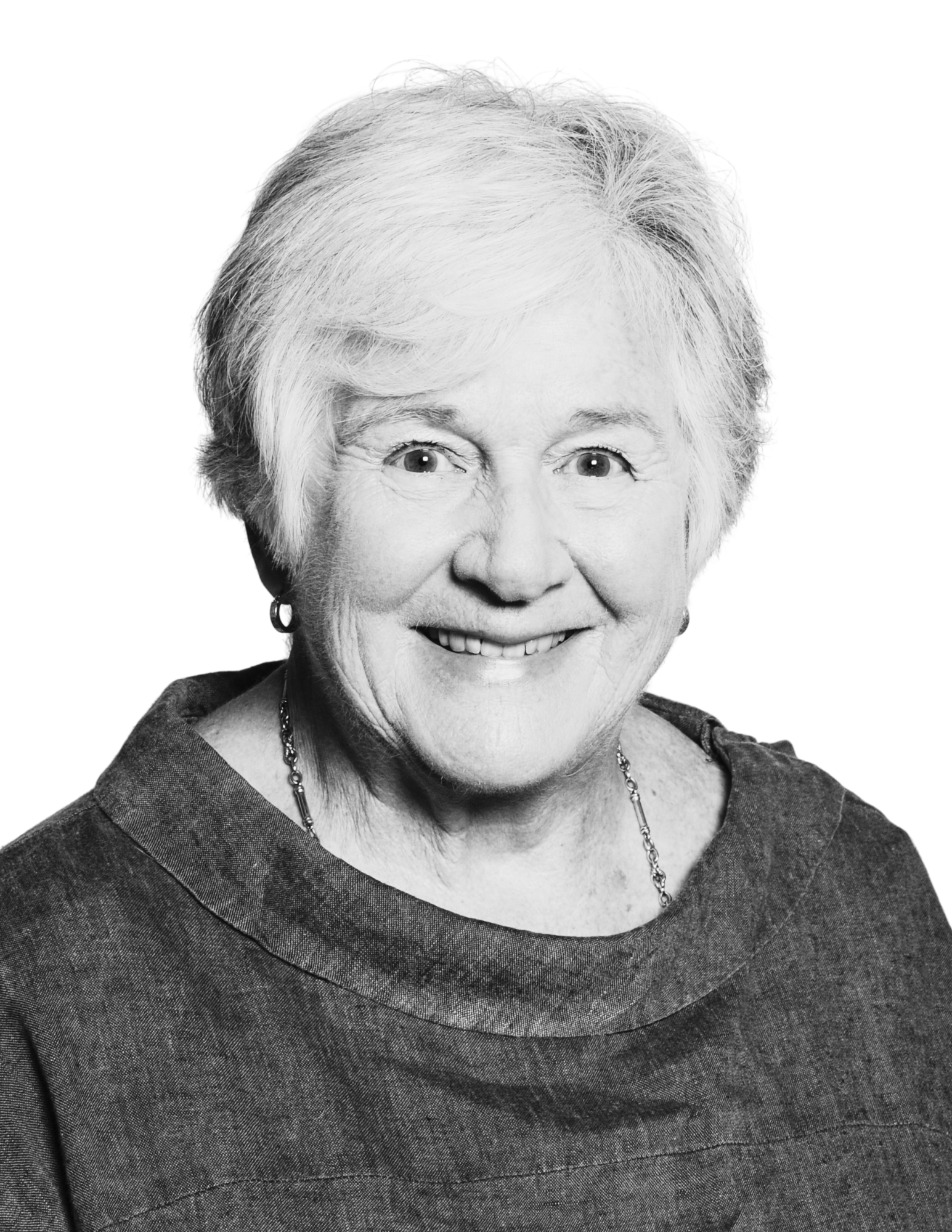 Black and white portrait of an elderly woman with short, light-coloured hair, smiling, wearing a dark top, earrings, and a necklace.