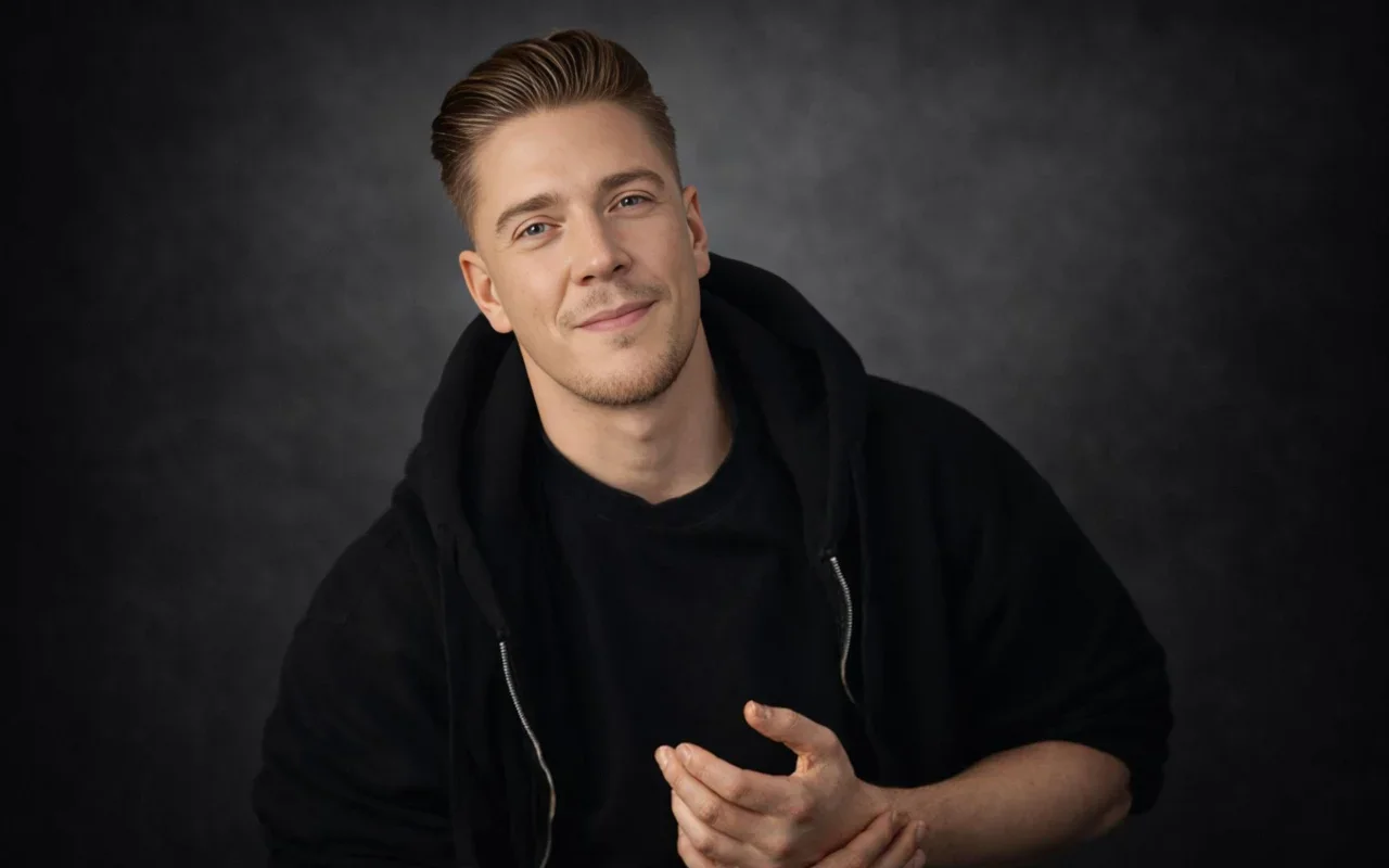 Portret van Dennis Bouwmeester, lachend en gekleed in een zwarte hoodie, tegen een donkere achtergrond.