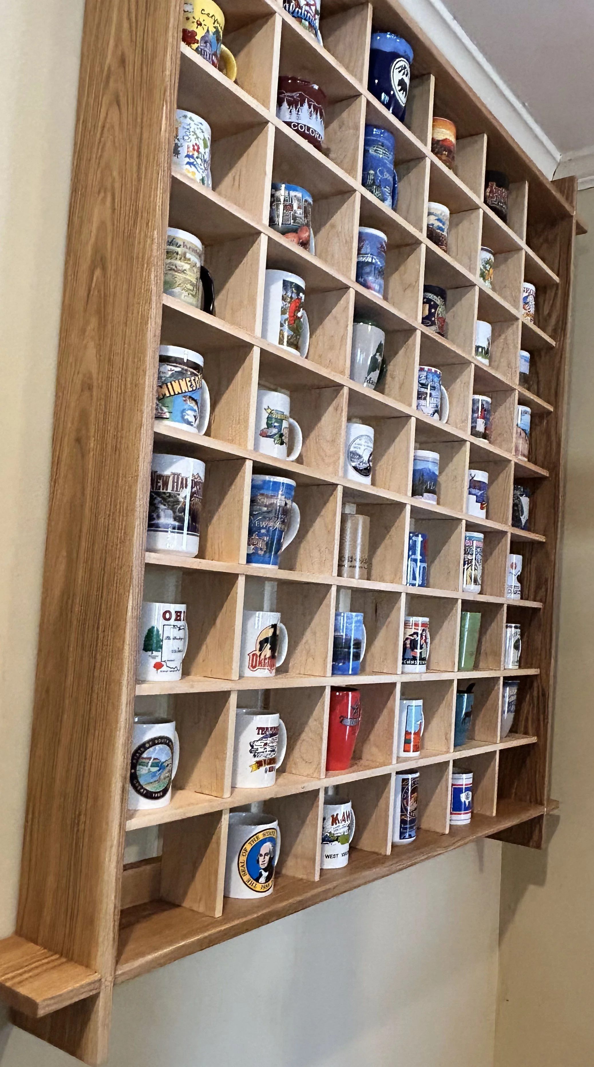 A wooden cubby shelf mounted on the wall with multiple individual compartments, each containing a souvenir mug with various colorful and scenic designs.
