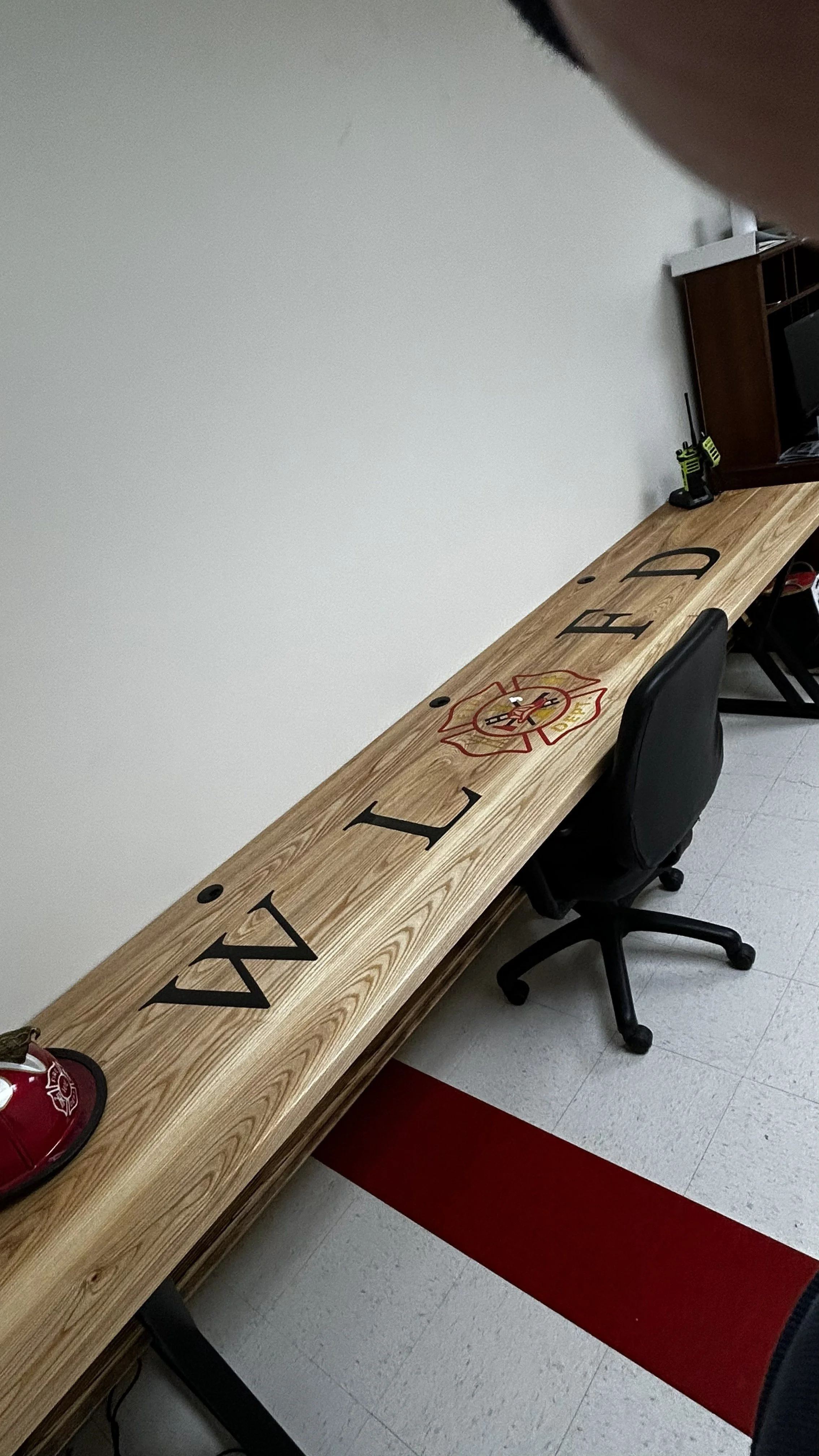 Wooden table with a fire department emblem and black letters, in a room with a black office chair, a fire helmet, a black and yellow power drill, and a wooden shelf.