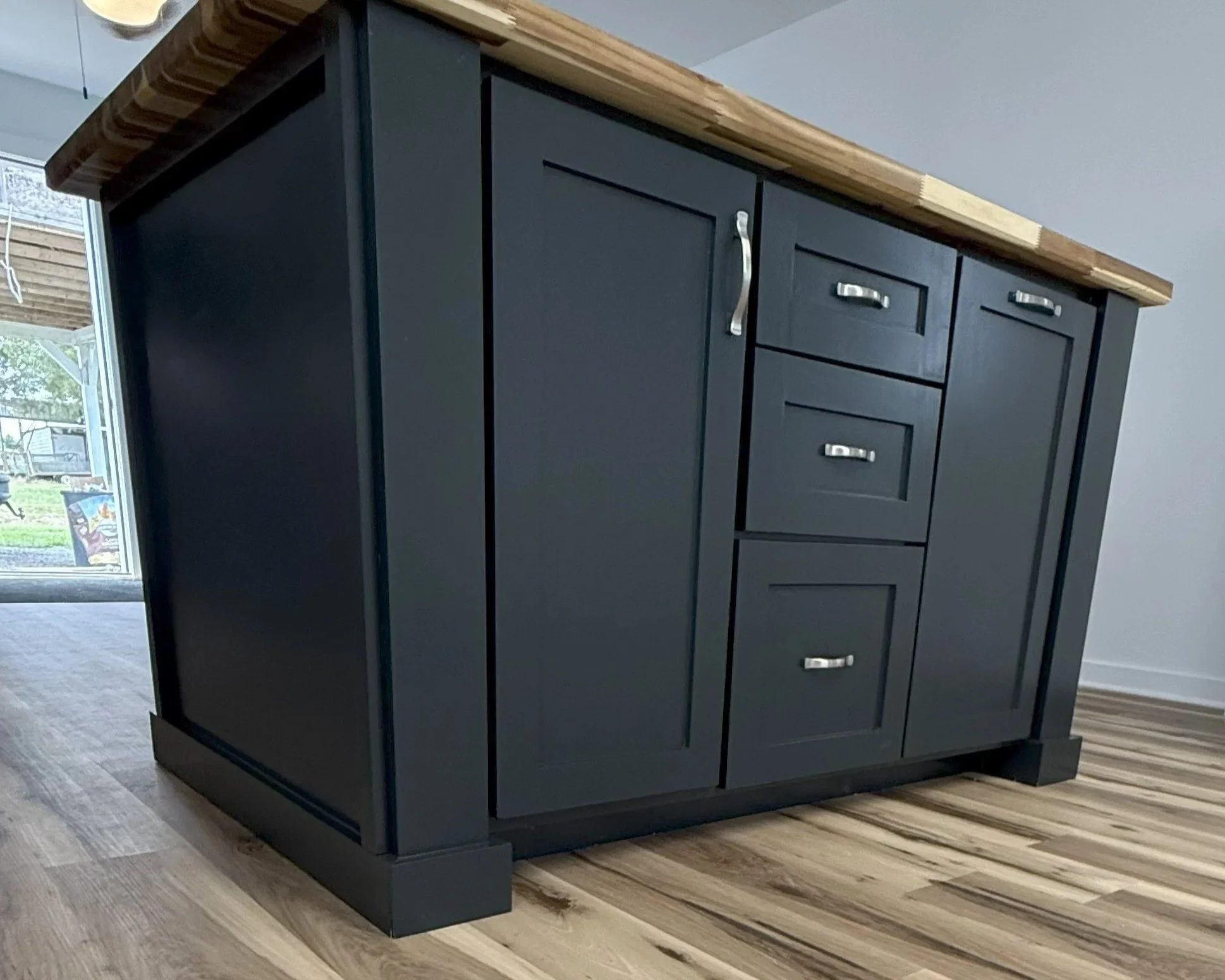 Black kitchen island with silver handles and a wooden countertop in a room with wooden floor and window in the background.
