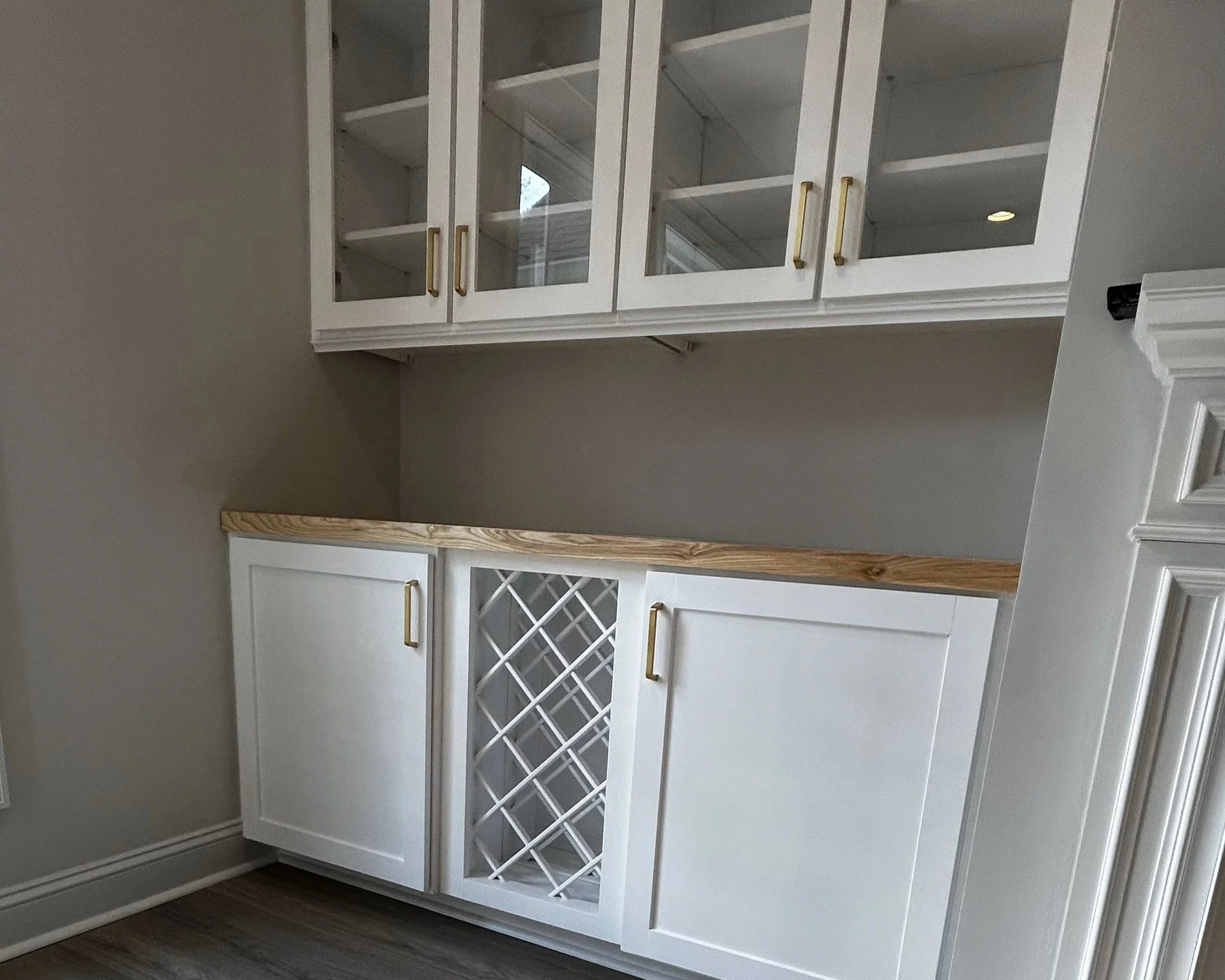 White built-in kitchen cabinets with glass-door upper cabinets with gold handles and white wine rack in the lower cabinet, adjacent to a staircase with white railing.