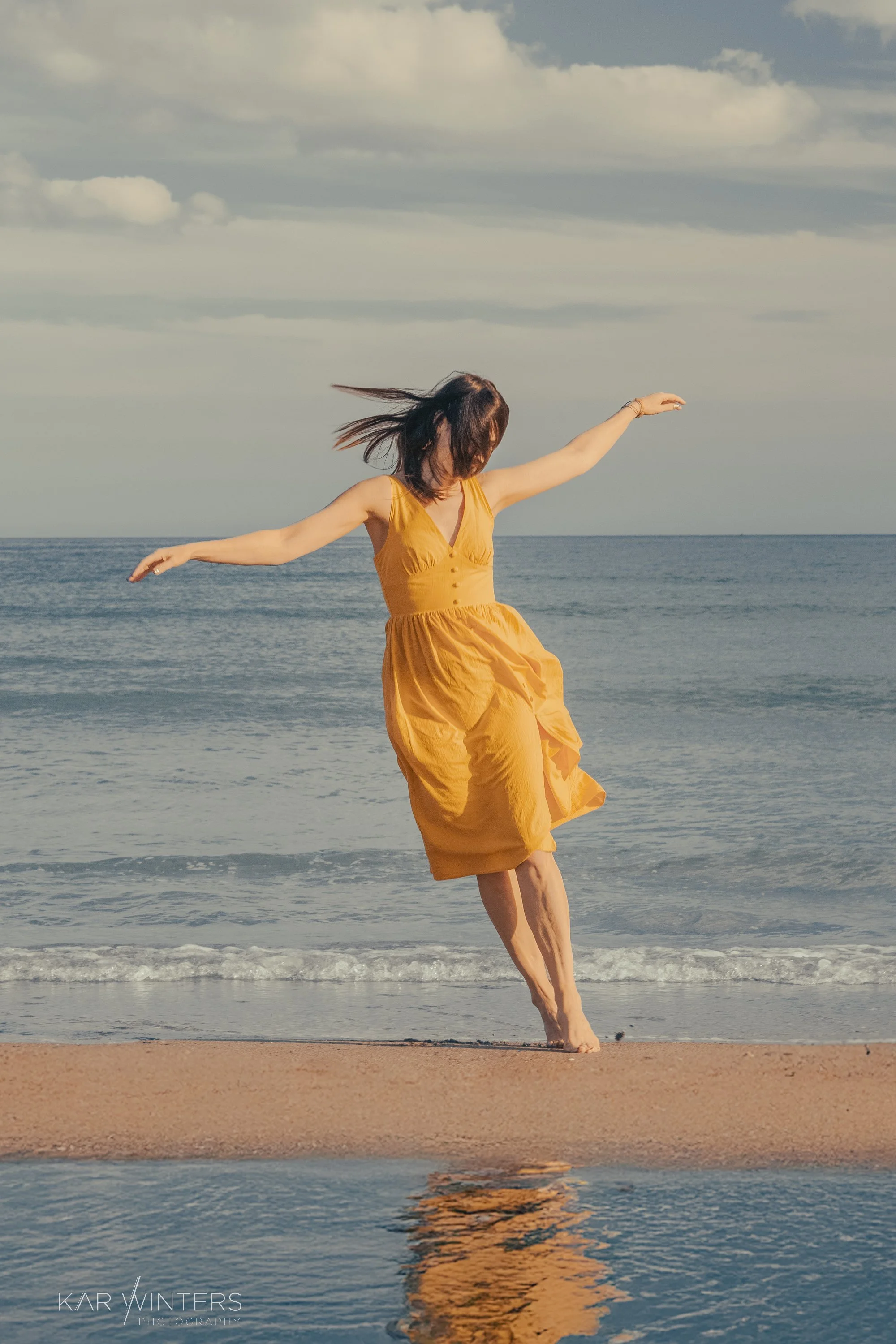 A woman in a yellow dress dancing on the beach near the water with her arms extended, wind blowing her hair, and cloudy sky above.