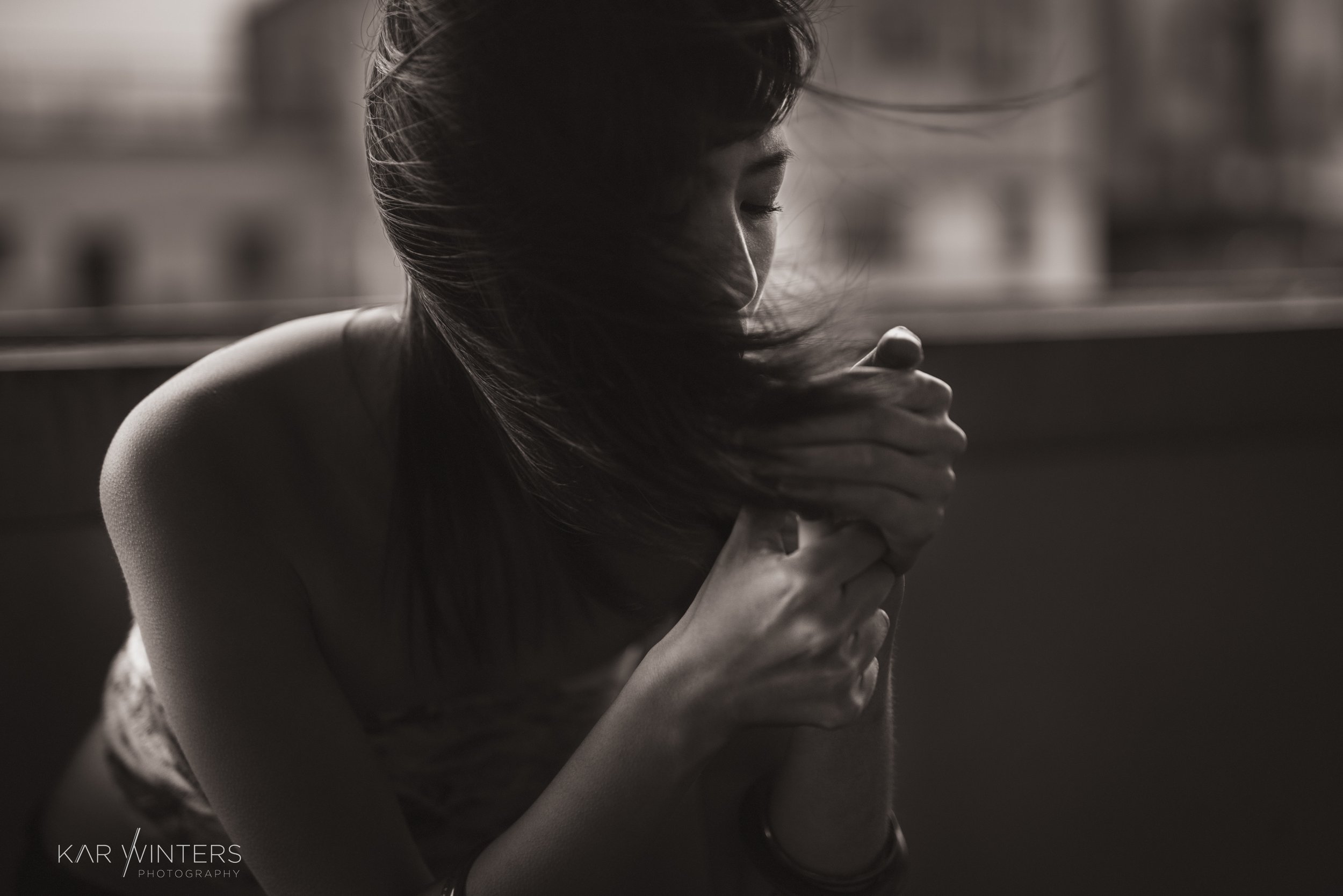 A woman with long hair, eyes closed, appears to be hugging herself or holding something close, in a contemplative or emotional moment with soft lighting and a blurred background.