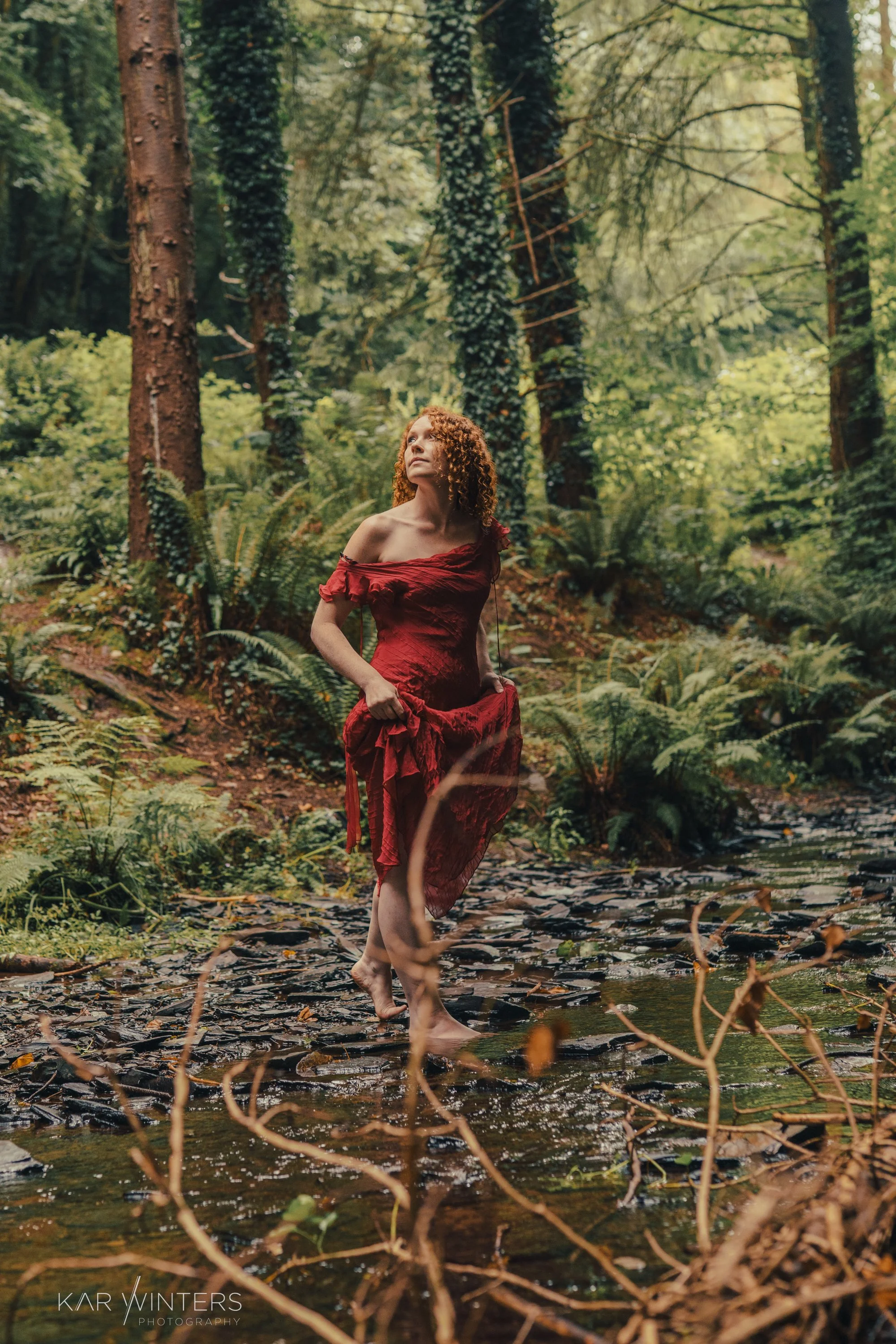 A woman with curly red hair wearing a red dress, walking barefoot through a shallow stream in a lush, green forest.