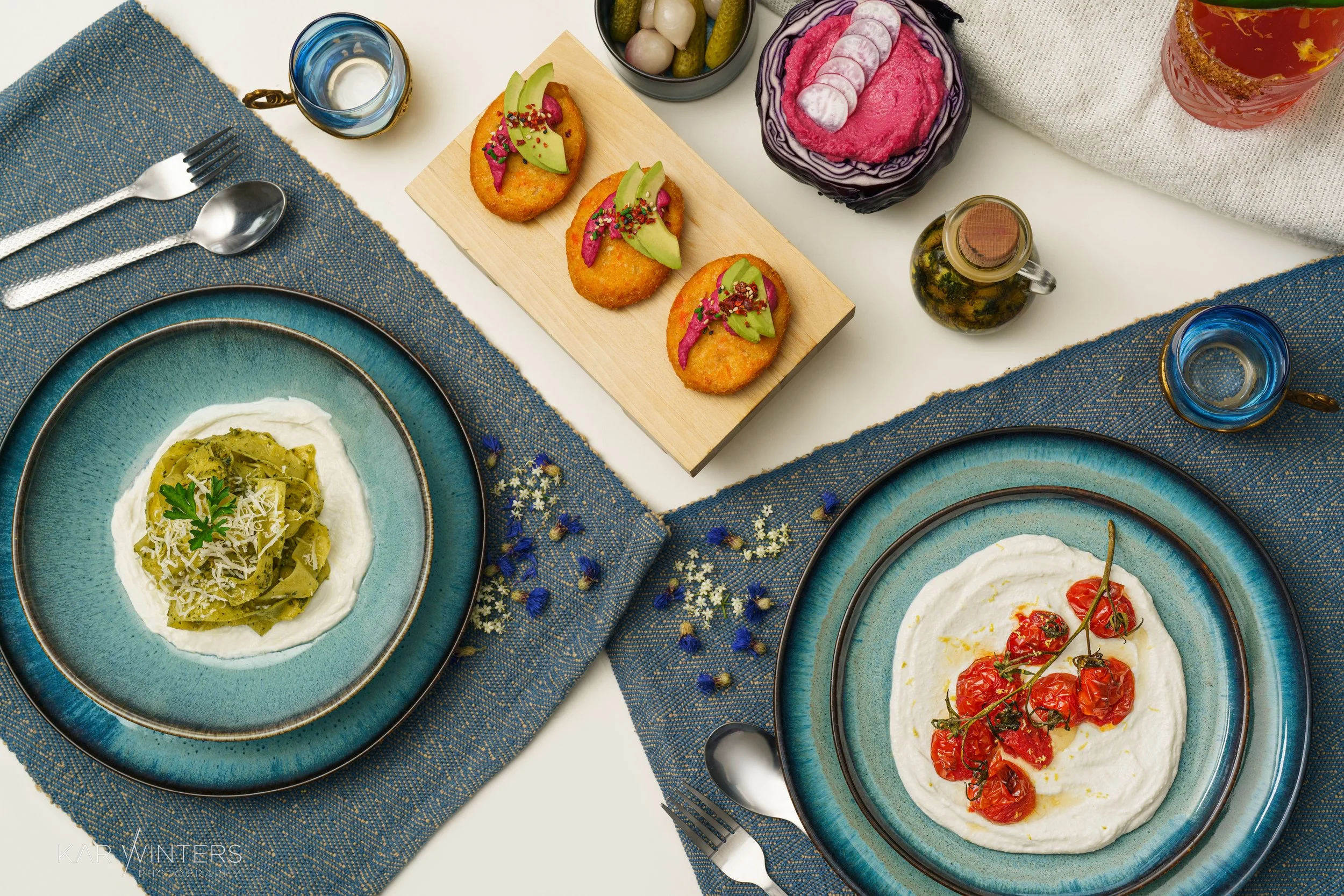 A top-down view of a dining table with two blue plates of pasta, a wooden serving tray with three breaded appetizers topped with avocado and sprouts, small bowls of pickles and pink dip, and glasses of water, salt, and pepper shakers, on a white tabl